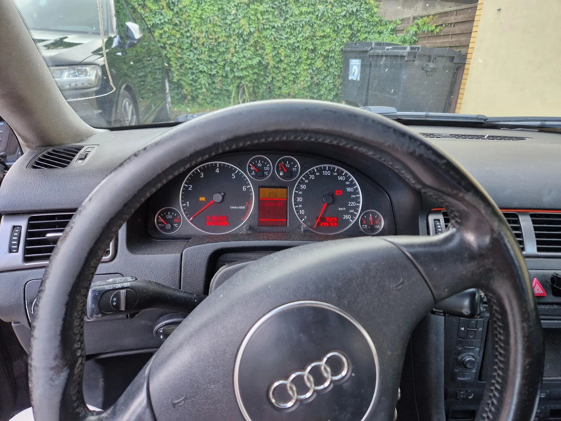 Widok z wnętrza samochodu marki Audi, skupiony na kierownicy i panelu instrumentów. Na desce rozdzielczej widoczne są analogowe wskaźniki prędkości i obrotów oraz czerwona kontrolka EPC. Na cyfrowym wyświetlaczu widoczny jest przebieg 295 705 km. Kierownica jest wykonana z czarnej skóry z logo Audi w centralnej części.