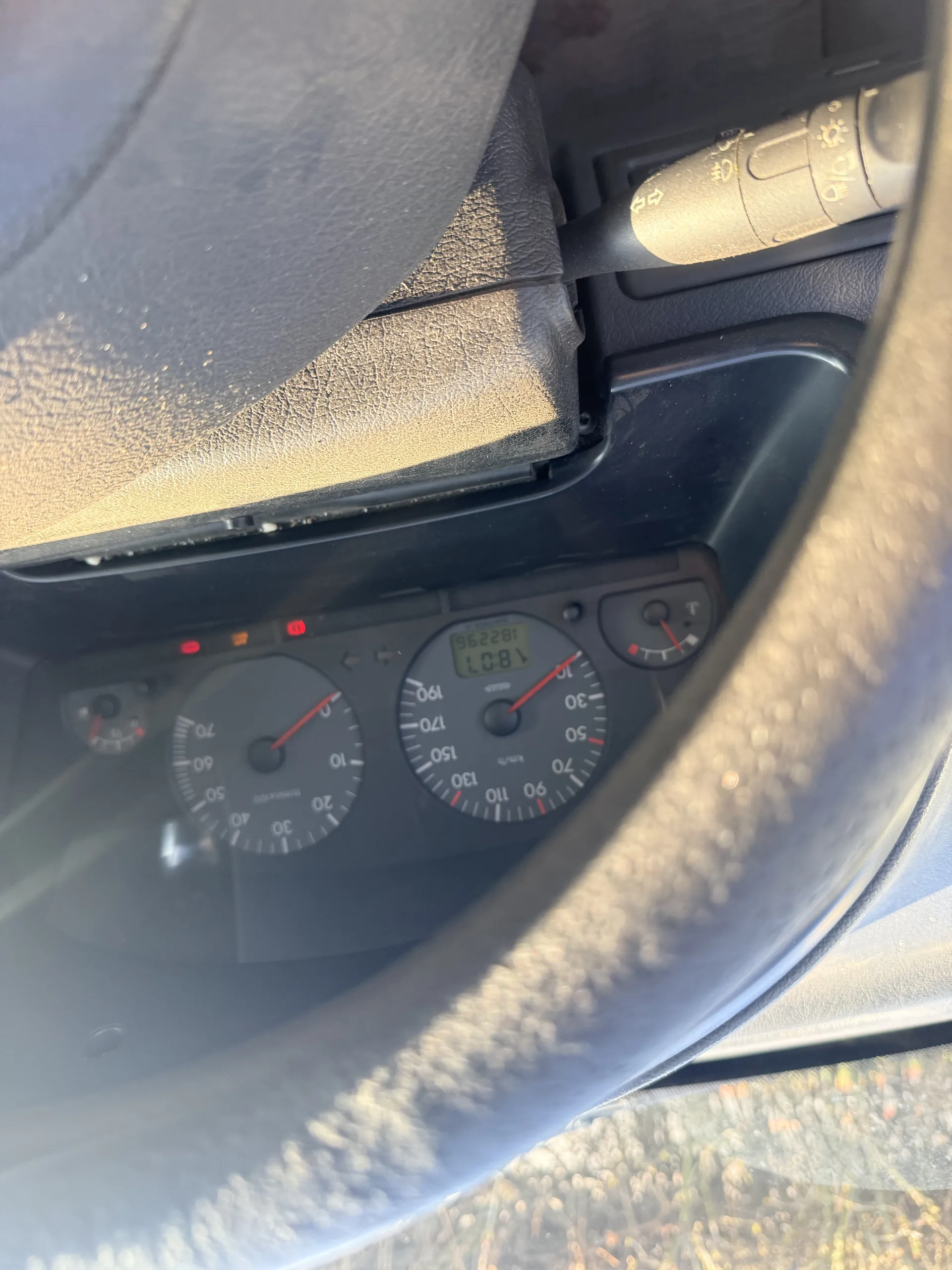 Deska rozdzielcza samochodu z prędkościomierzem (skala do 190 km/h), obrotomierzem (skala do 70), wyświetlaczem cyfrowym pokazującym godzinę 18:07 i przebieg 182296. Widoczne są czerwone lampki ostrzegawcze. Fragment kierownicy po lewej stronie, po prawej kolumna kierownicy z przyciskami sterującymi światłami. W tle przez okno widoczna trawa i światło słoneczne.