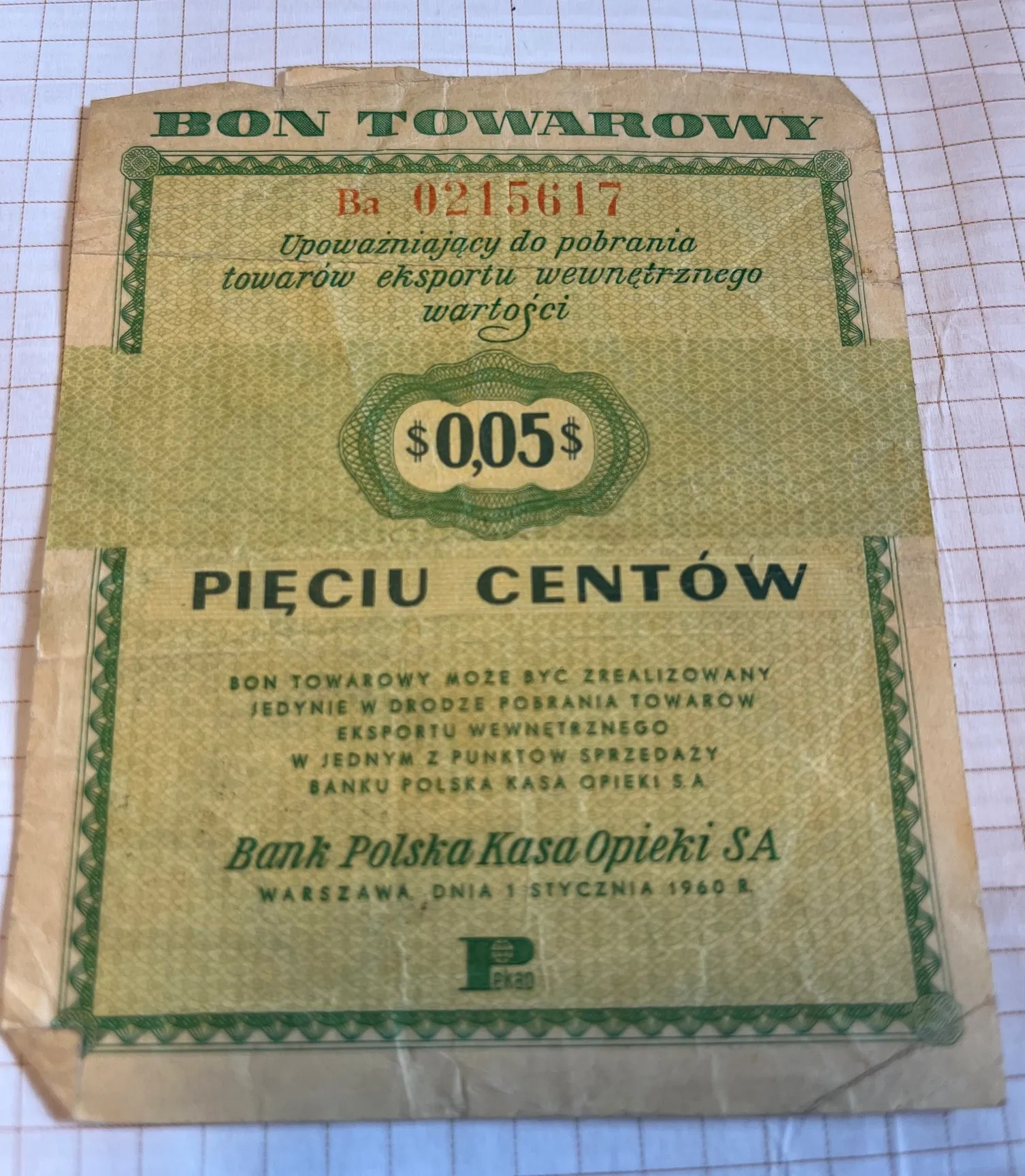 Stary, pożółkły bon towarowy z zielonymi zdobieniami w formie giloszu. Na górze widnieje napis 'BON TOWAROWY' oraz numer 'Ba 0215617'. Poniżej znajduje się tekst: 'Upoważniający do pobrania towarów eksportu wewnętrznego wartości'. W centralnej części, w ozdobnej ramce, widnieje kwota '$0,05$', a pod nią napis 'PIĘCIU CENTÓW'. W dolnej części znajduje się informacja o zasadach realizacji bonu, nazwa instytucji 'Bank Polska Kasa Opieki SA' oraz data 'WARSZAWA DNIA 1 STYCZNIA 1960 R.'. Na samym dole widoczne jest małe logo 'ekad'.
