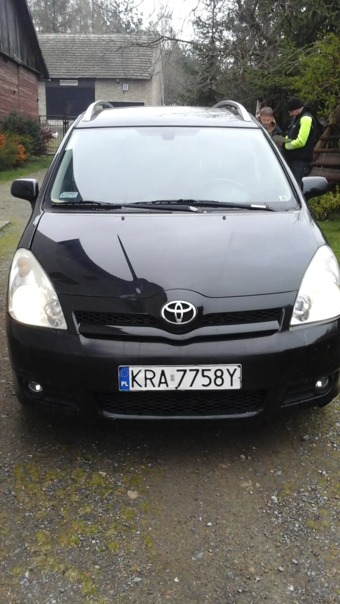 Czarny samochód marki Toyota z tablicą rejestracyjną KRA 7758Y. Przy samochodzie stoją dwie osoby, jedna w jasnozielonej kurtce. Tło: dom z dachem krytym dachówką, drzewa i krzewy. Samochód zaparkowany na żwirowej powierzchni.