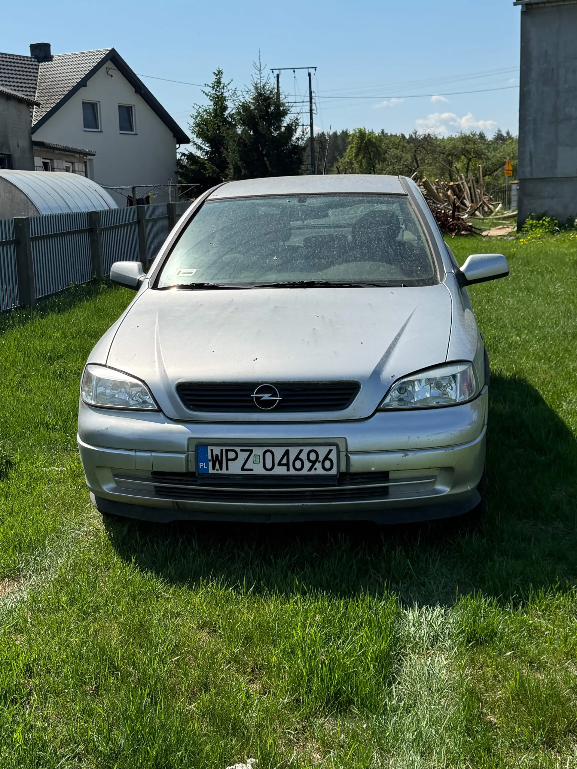 Srebrny samochód marki Opel z tablicą rejestracyjną WPZ 04696 stojący na zielonej trawie. W tle dom z szarą dachówką, metalowy płot, drzewa, słup energetyczny oraz jasne niebo. Po prawej stronie widoczna betonowa konstrukcja i stos drewna.