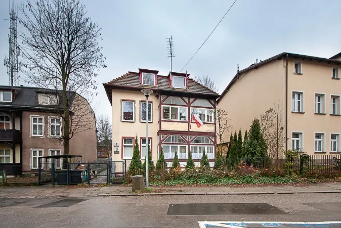 Zdjęcie przedstawiające trzy budynki mieszkalne wzdłuż ulicy. Po lewej znajduje się ciemnobrązowy dom z białymi obramowaniami okien. W centrum stoi dwupiętrowy dom o jasnej elewacji z elementami konstrukcji szkieletowej (mur pruski) na wyższych kondygnacjach, z czerwoną flagą zawieszoną na balkonie. Po prawej widoczny jest beżowy, prosty budynek wielorodzinny. Przed domami znajduje się chodnik, niska roślinność, krzewy oraz metalowe ogrodzenie. W tle po lewej stronie widoczny jest wysoki maszt telekomunikacyjny.