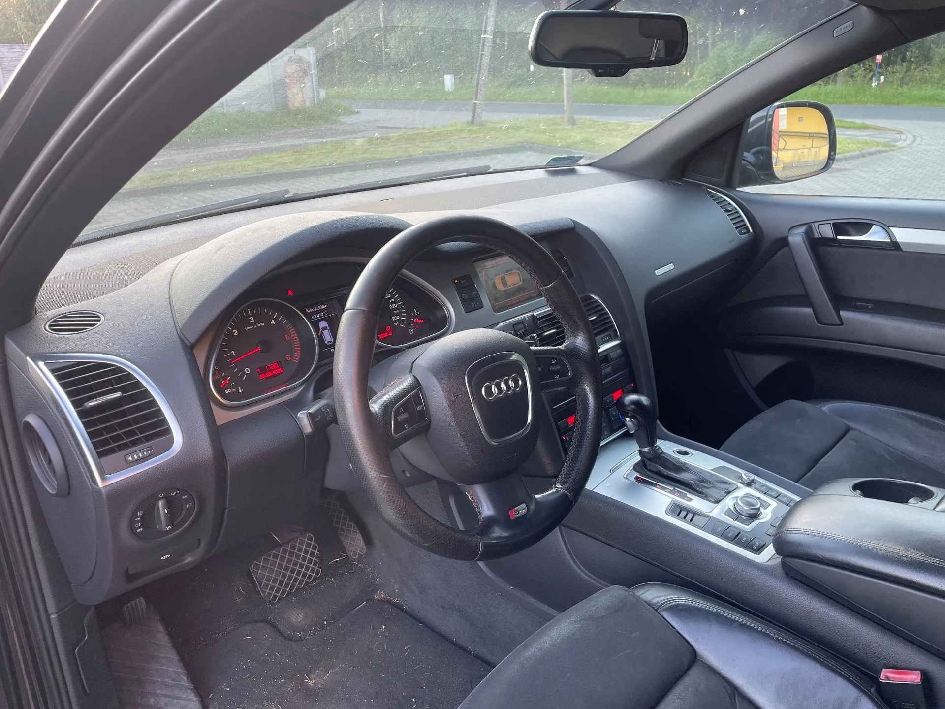 Widok z wnętrza samochodu marki Audi, ukazujący deskę rozdzielczą, kierownicę z logo Audi oraz konsolę środkową z lewarkiem zmiany biegów. Na panelu instrumentów widoczne są wskaźniki prędkości i obrotów oraz mały wyświetlacz z czerwonym tekstem. Przez przednią szybę widoczny jest fragment drogi i zieleni.