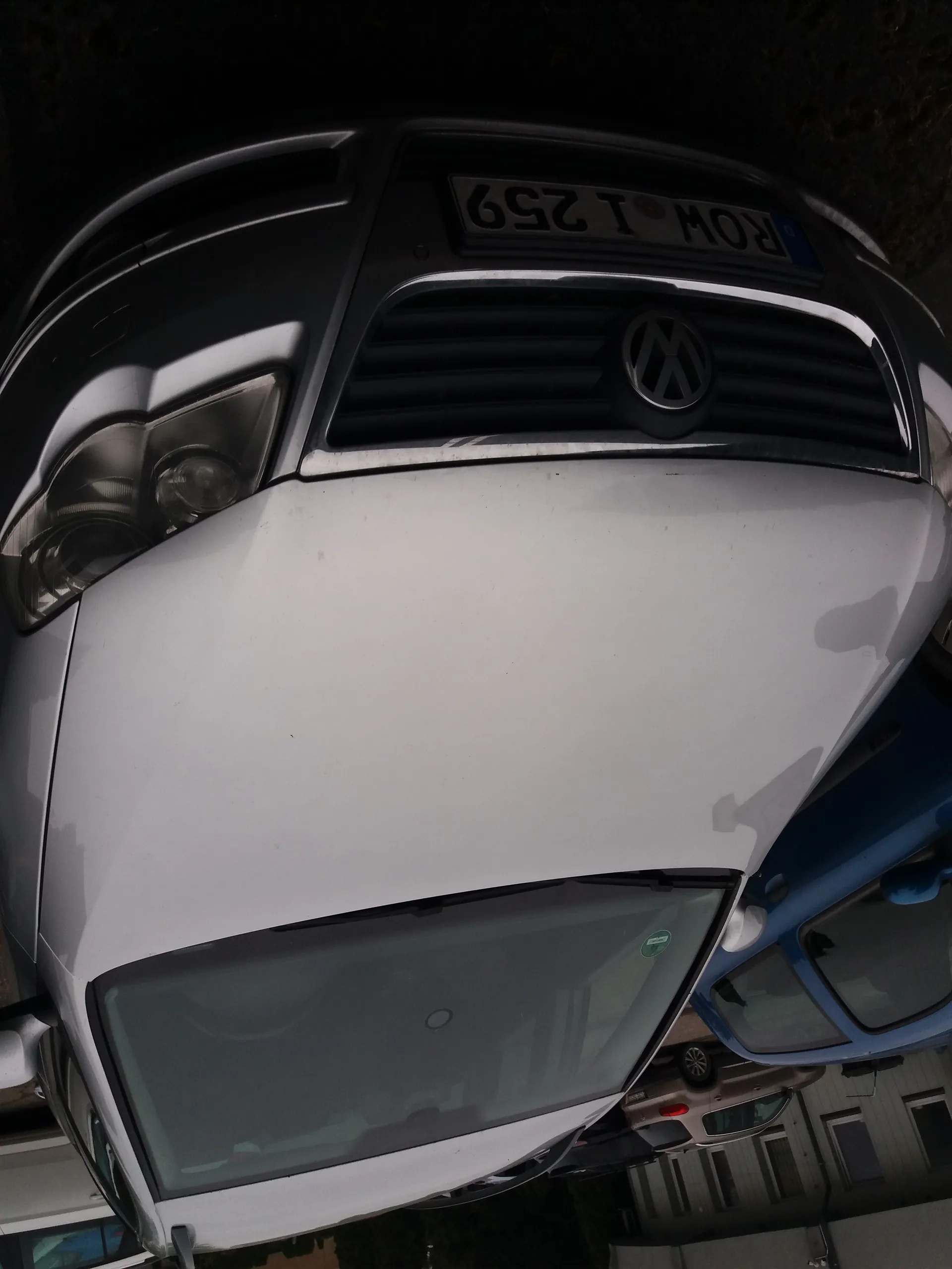 Biały samochód marki Volkswagen z przodu, z widocznym logo na atrapie chłodnicy i tablicą rejestracyjną ROW I 259. W tle niebieski samochód oraz budynki mieszkalne.