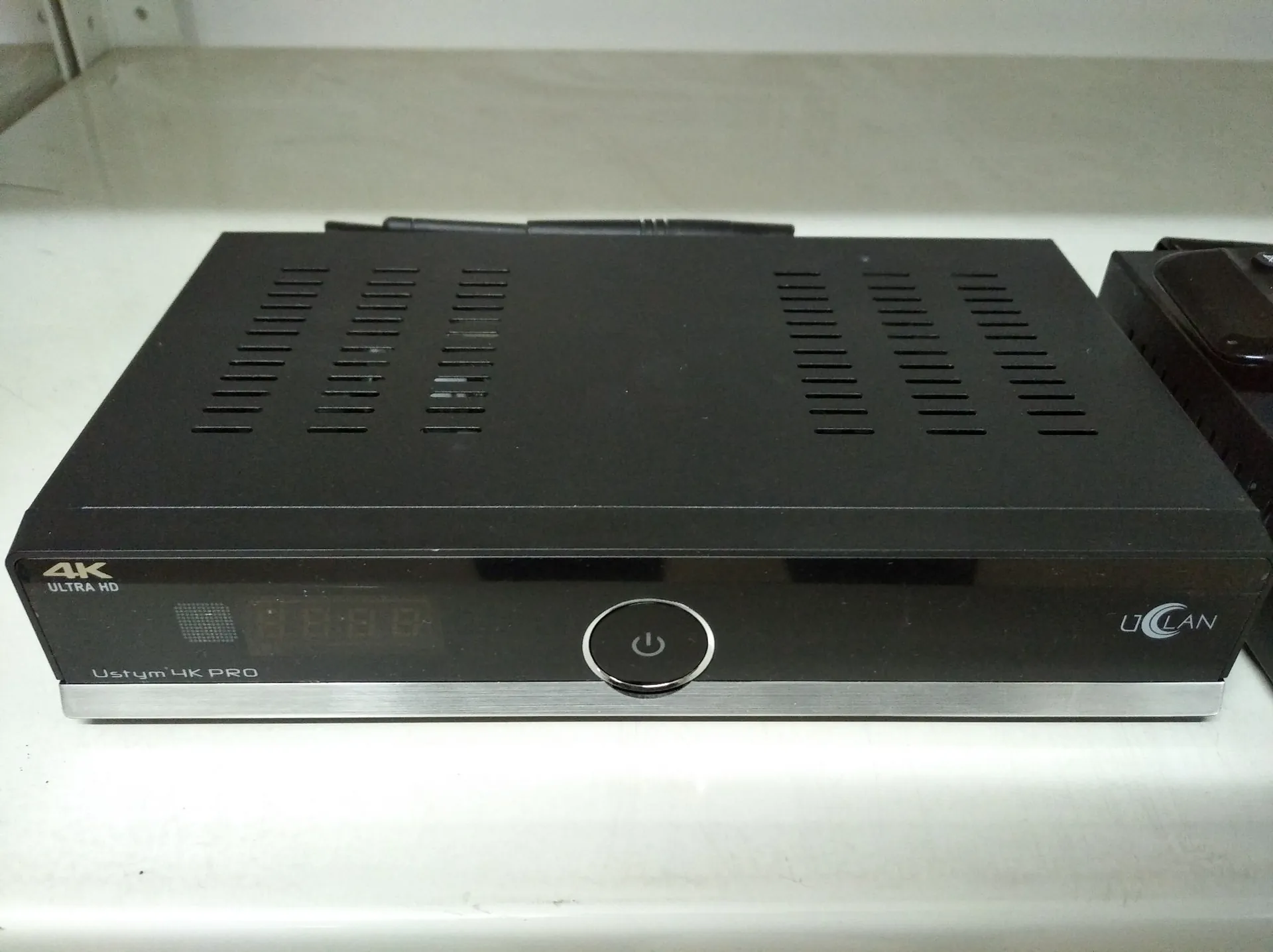 Czarny dekoder lub odtwarzacz multimedialny o prostokątnym kształcie, leżący na białej powierzchni. Na przednim panelu widnieje napis '4K ULTRA HD' po lewej stronie, okrągły przycisk zasilania na środku oraz napis 'UCLAN' po prawej stronie. Na dolnej srebrnej ramce znajduje się napis 'Ustym 4K PRO'. Górna obudowa urządzenia posiada otwory wentylacyjne w formie pionowych szczelin.