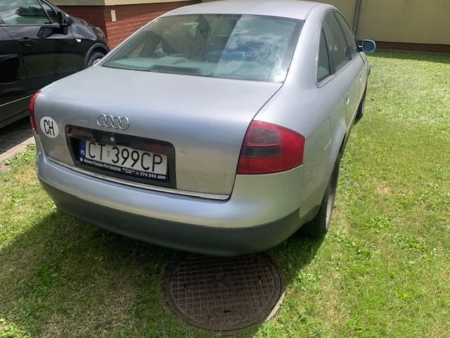 Srebrny samochód marki Audi widziany od tyłu, zaparkowany na trawie. Na klapie bagażnika widoczne logo Audi oraz tablica rejestracyjna o numerze CT 399CP. Po lewej stronie tablicy znajduje się owalna naklejka z napisem CH.