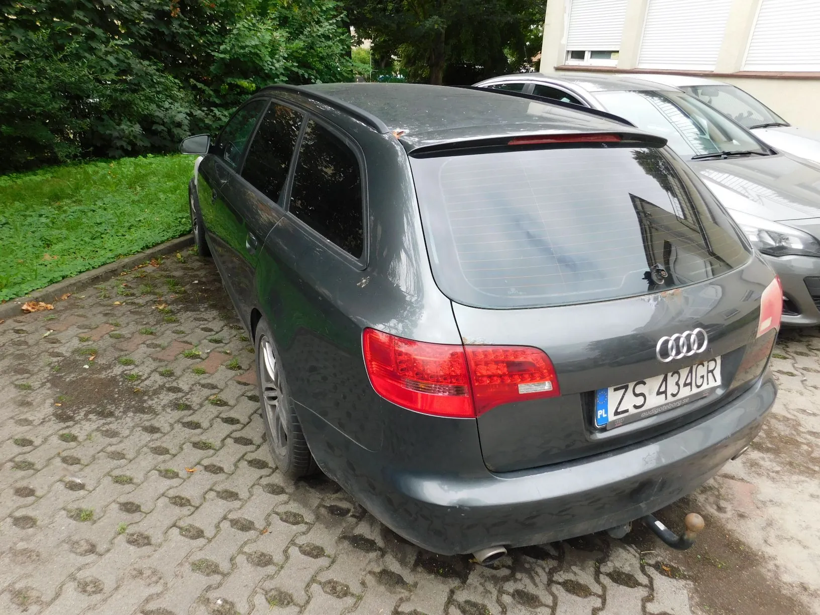 Szary samochód marki Audi w nadwoziu kombi zaparkowany tyłem na kostce brukowej. Widoczna jest tylna klapa z logo Audi oraz polska tablica rejestracyjna o numerze ZS 434GR. Samochód znajduje się na tle zielonych krzewów i fragmentu jasnego budynku.