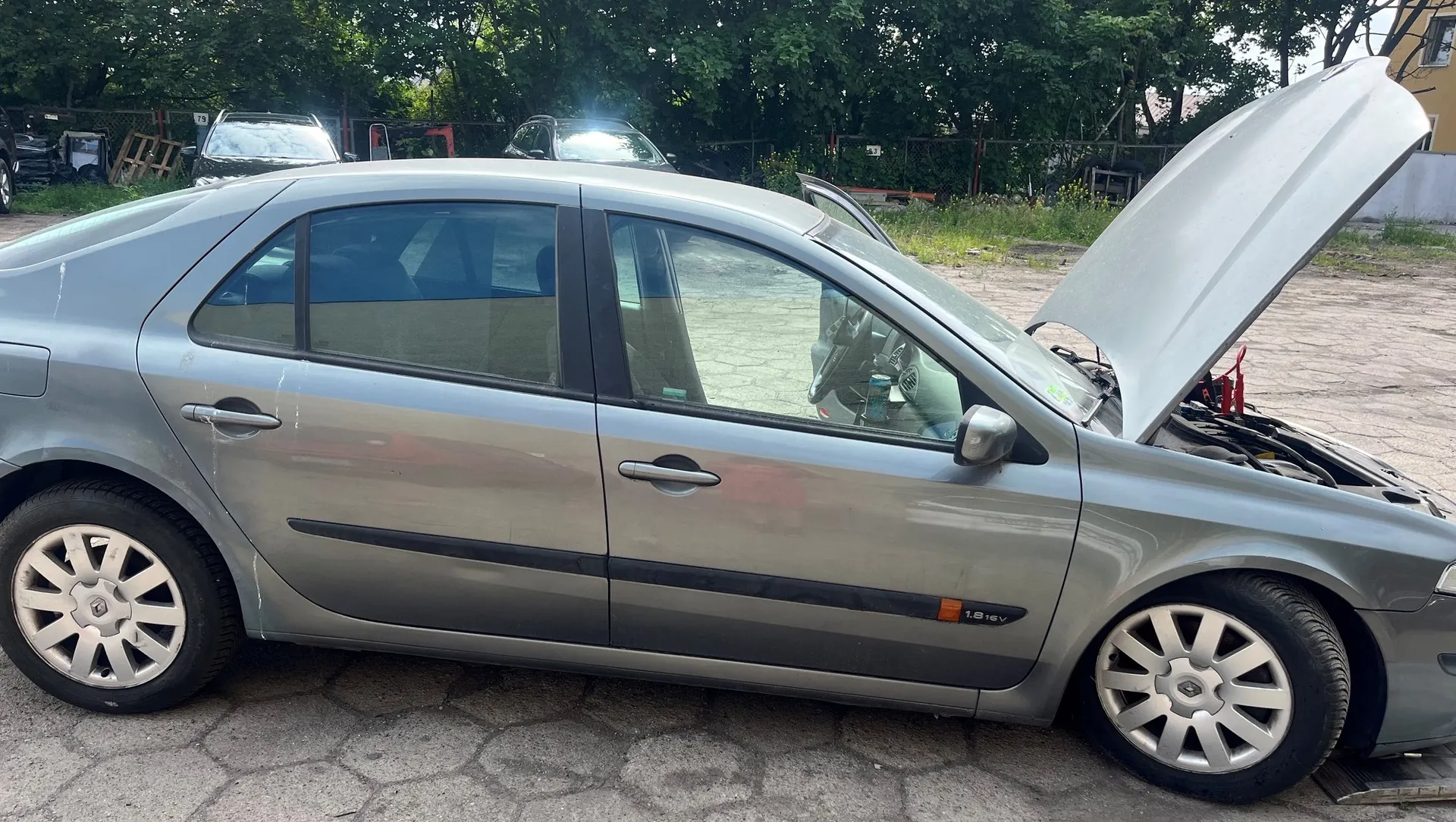 Srebrny samochód osobowy w nadwoziu hatchback zaparkowany na betonowym placu. Maski samochodu jest otwarta, odsłaniając przedział silnika. Na przednich drzwiach widoczny jest napis '1.8 16V'. W tle znajdują się inne zaparkowane samochody oraz zielone drzewa.