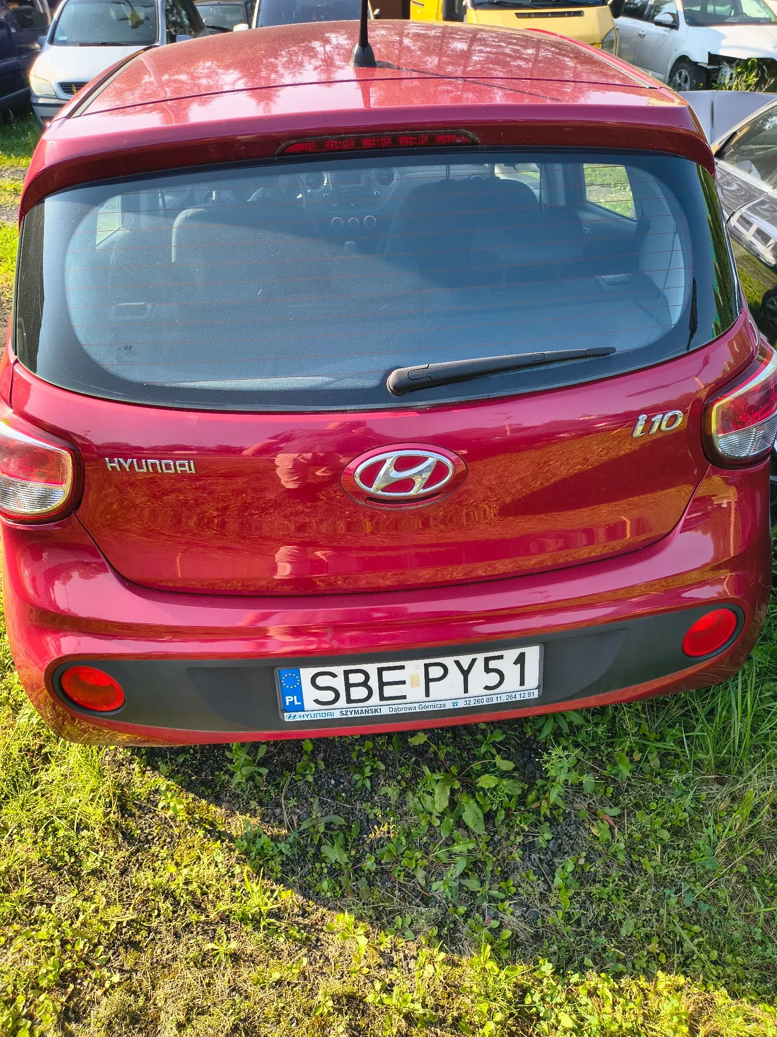 Tylny widok czerwonego samochodu marki Hyundai i10. Na klapie bagażnika widoczne jest logo Hyundai oraz napisy 'HYUNDAI' po lewej i 'i10' po prawej stronie. Samochód posiada polską tablicę rejestracyjną o numerze SBE PY51. Poniżej tablicy znajduje się ramka z napisem 'HYUNDAI SZYMARSKI Dobczyce Gorzów Wlkp. 32 260-03 11, 264 12 01'. Auto stoi na trawiastym podłożu, w tle widoczne są inne samochody.