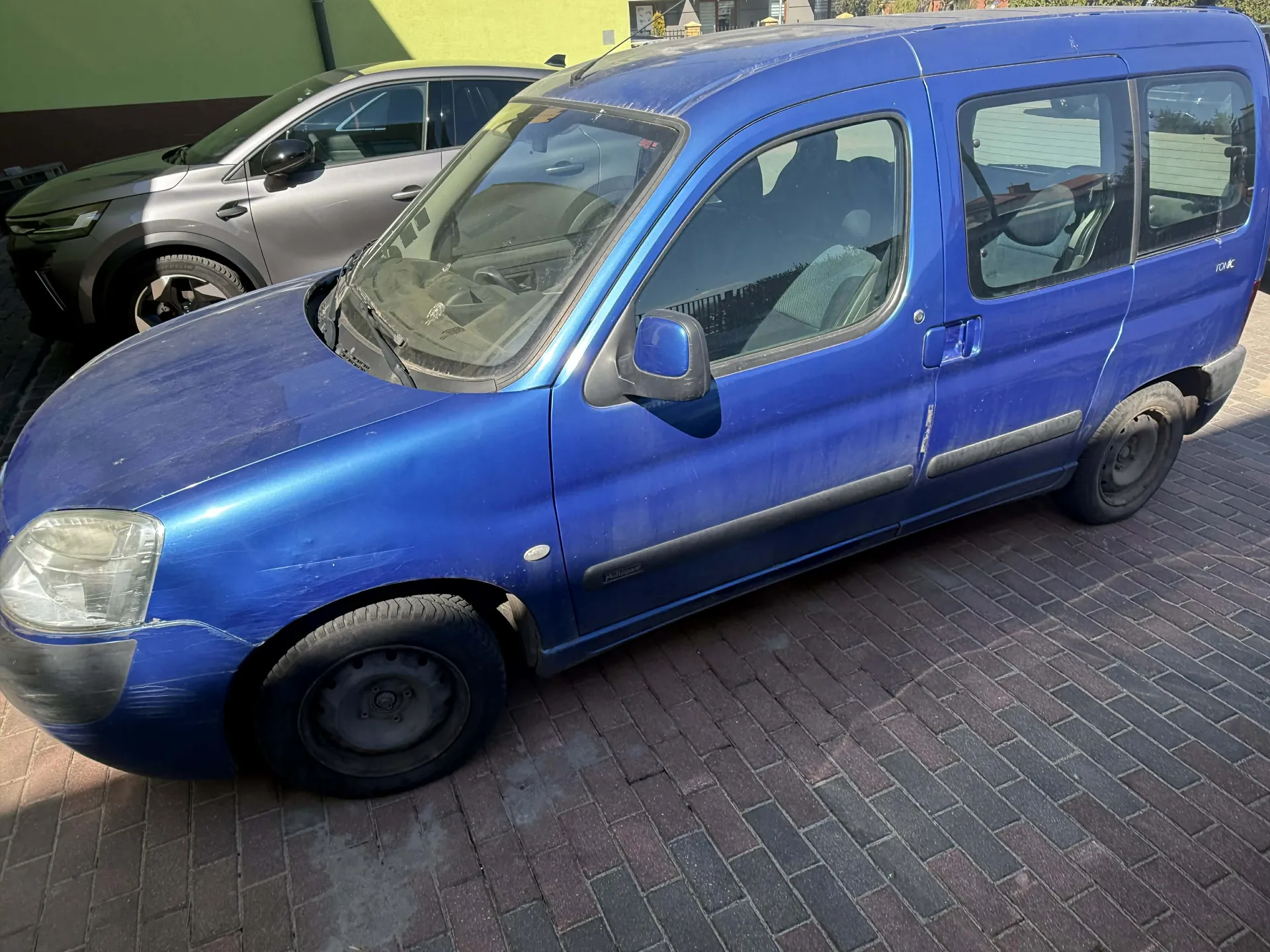Niebieski furgonetka zaparkowana na brukowanej powierzchni. Po lewej stronie widoczny szary samochód osobowy. Furgonetka ma zarysowane uszkodzenia na przednim błotniku, brudną karoserię i czarne felgi. W tle zielona ściana budynku oraz fragmenty innych obiektów. Okna furgonetki są przejrzyste, widać wnętrze z siedzeniami i kierownicą.