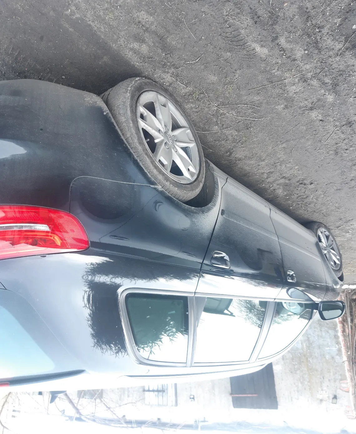 Czarny samochód osobowy typu sedan, widok z tyłu i boku. Widoczne czerwone światło tylnego reflektora, srebrna felga aluminiowa oraz fragment ciemnej nawierzchni drogi. W tle budynek z oknami i drzwiami oraz drzewa bez liści.