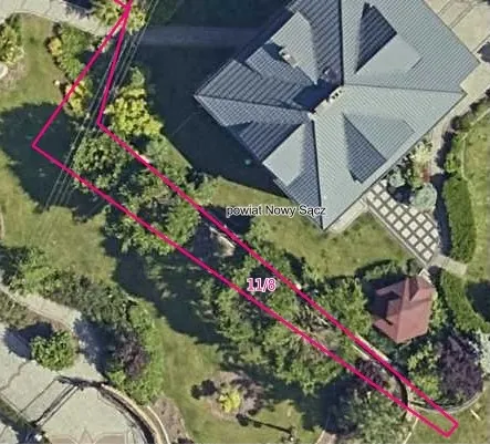 Zdjęcie satelitarne przedstawiające fragment posesji z domem jednorodzinnym o szarym, wielospadowym dachu. Na obrazie naniesiono różową linię wyznaczającą granicę działki oraz tekst: 'powiat Nowy Sącz' i numer '11/8'. Wokół domu znajduje się zielony trawnik, drzewa, krzewy oraz utwardzona ścieżka z kostki brukowej.