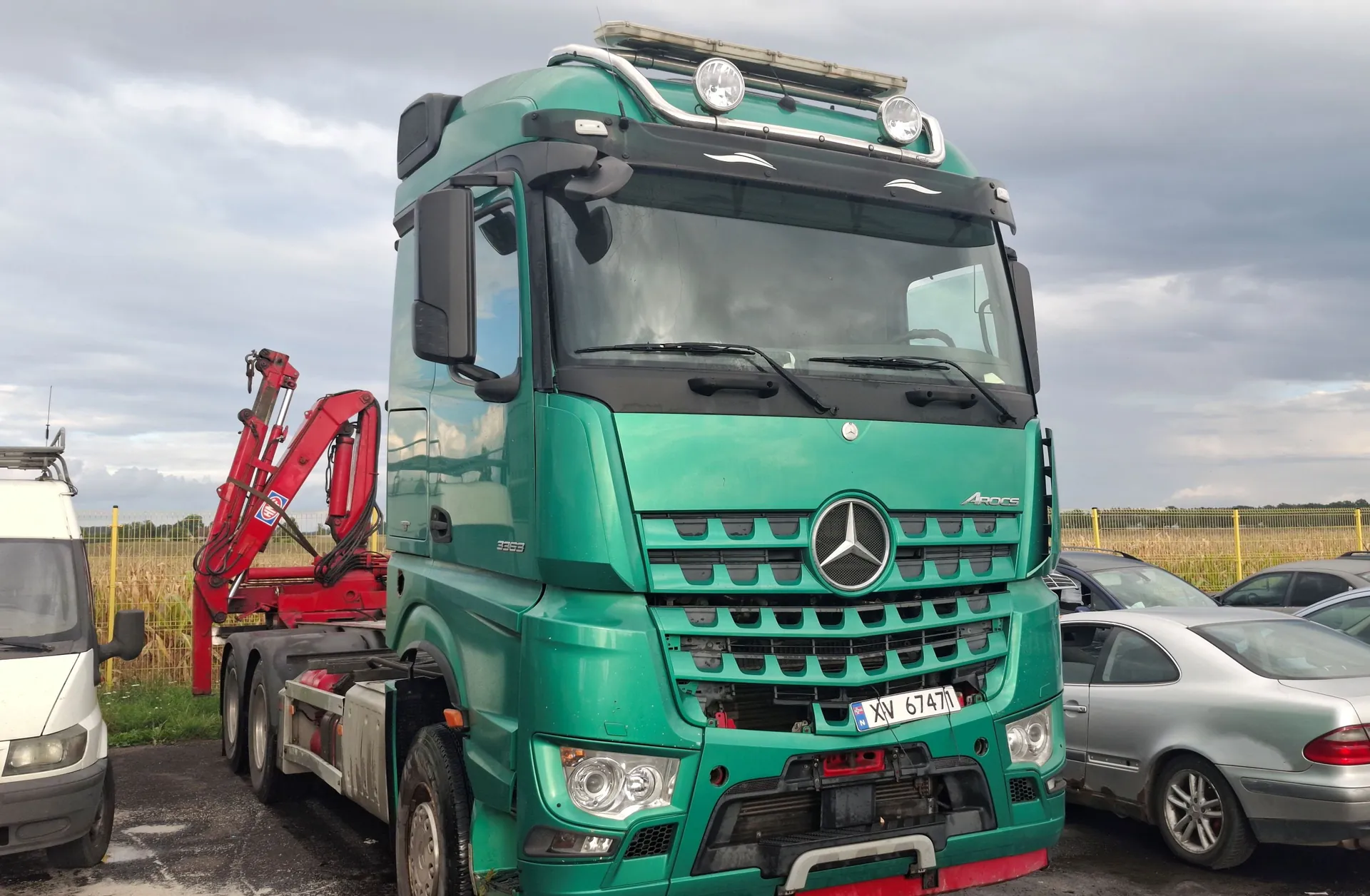 Zielona ciężarówka marki Mercedes-Benz Actros z zamontowanym czerwonym żurawiem ładunkowym z tyłu. Pojazd stoi na parkingu pod zachmurzonym niebem. Na przednim zderzaku widoczna jest tablica rejestracyjna o numerze XV 6747N. Obok ciężarówki zaparkowane są inne samochody, w tym biały furgon po lewej i srebrny samochód osobowy po prawej.