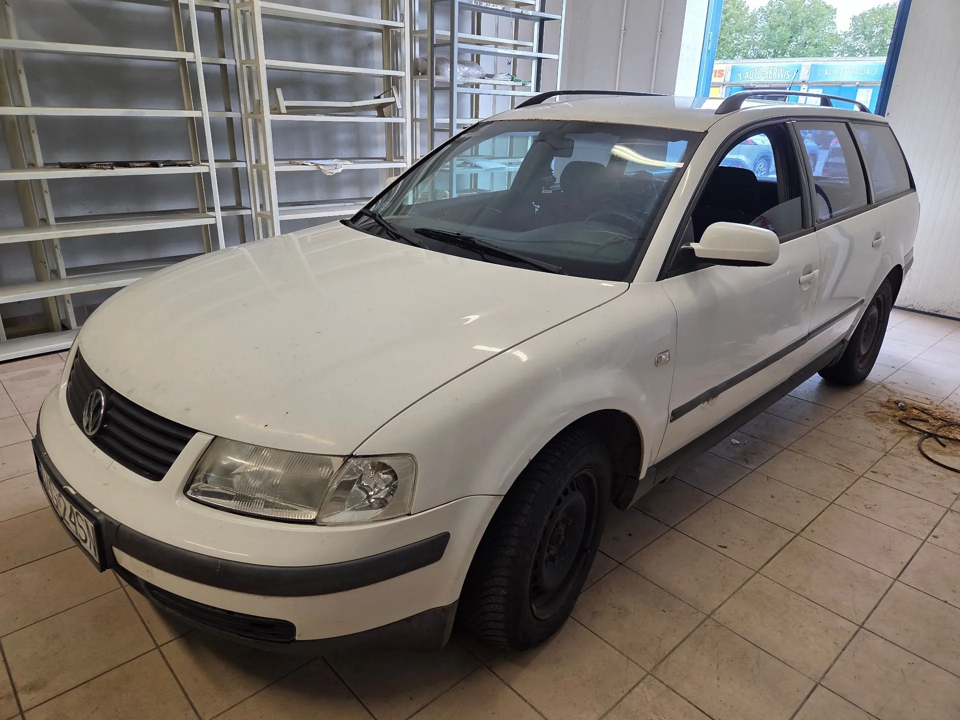 Biały samochód osobowy typu kombi marki Volkswagen zaparkowany wewnątrz pomieszczenia przypominającego warsztat lub magazyn. Pojazd stoi na płytkach ceramicznych, a w tle widoczne są białe, metalowe regały magazynowe. Samochód jest ustawiony pod kątem, ukazując przód i prawy bok nadwozia.