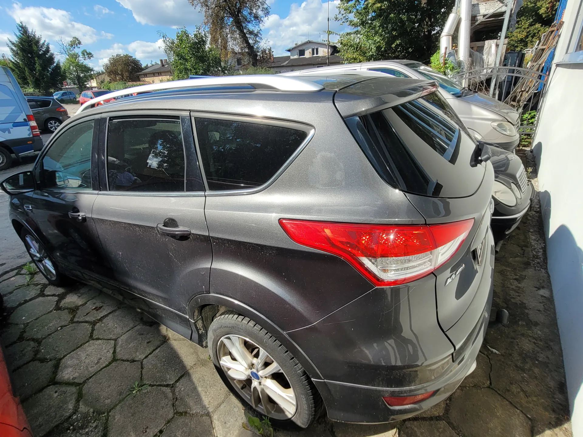 Srebrny SUV zaparkowany na brukowanej powierzchni, widok z tyłu i boku. Pojazd ma dachowy bagażnik, przyciemniane szyby i czerwone światła tylne. W tle inne samochody, drzewa, budynki mieszkalne oraz niebo z chmurami. Na tylnej części pojazdu widoczny niewyraźny napis. Podłoga z kamiennych płyt, po prawej stronie biała ściana budynku.