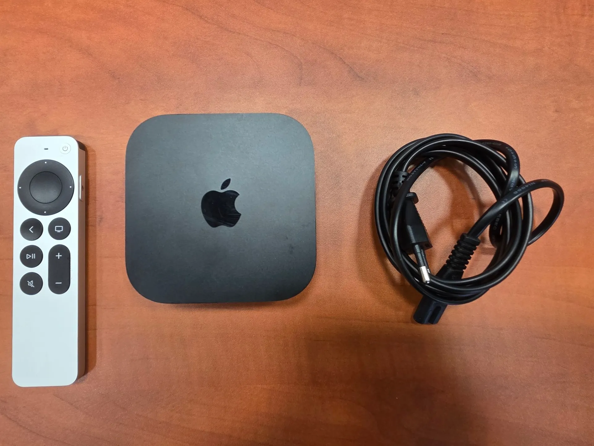 Na brązowym, drewnianym blacie leżą trzy przedmioty: biały pilot do Apple TV z czarnymi przyciskami, czarna kwadratowa przystawka Apple TV z logo jabłka na górnej obudowie oraz zwinięty czarny kabel zasilający.