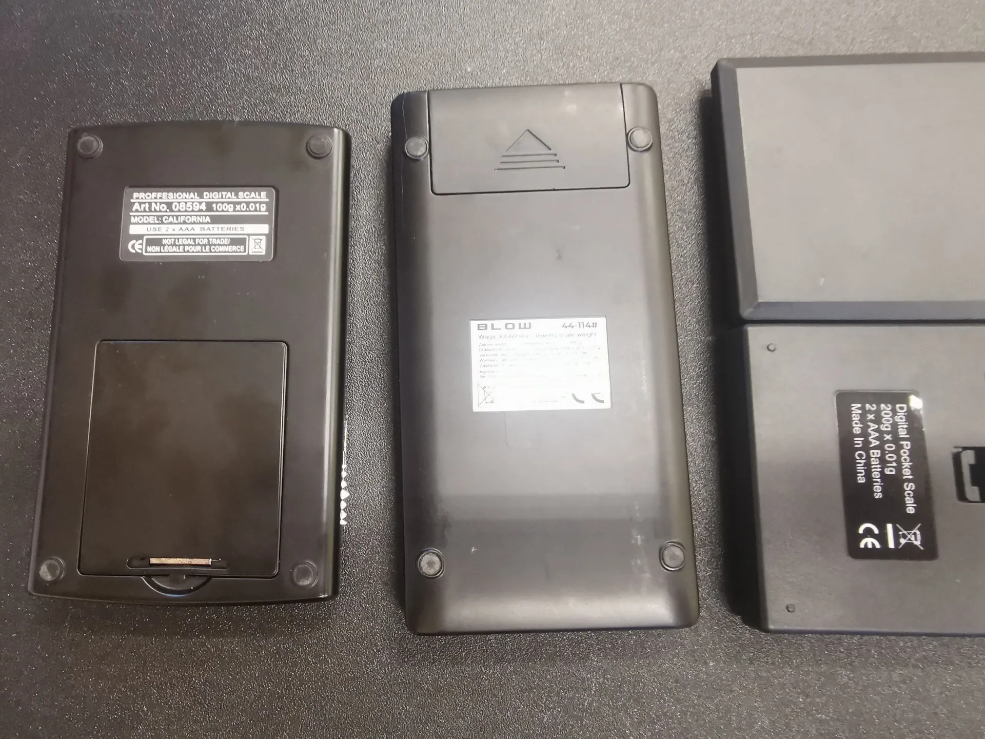 Zdjęcie trzech czarnych, prostokątnych urządzeń elektronicznych leżących obok siebie na szarej powierzchni. Pierwsze urządzenie od lewej posiada etykietę z tekstem: 'PROFFESIONAL DIGITAL SCALE Art No. 08594 100g x0.01g MODEL: CALIFORNIA'. Środkowe urządzenie ma logo 'BLOW' oraz numer '44-114#'. Trzecie urządzenie po prawej posiada etykietę z napisem: 'Digital Pocket Scale 200g x 0.01g 2 x AAA Batteries Made in China' oraz znak CE.
