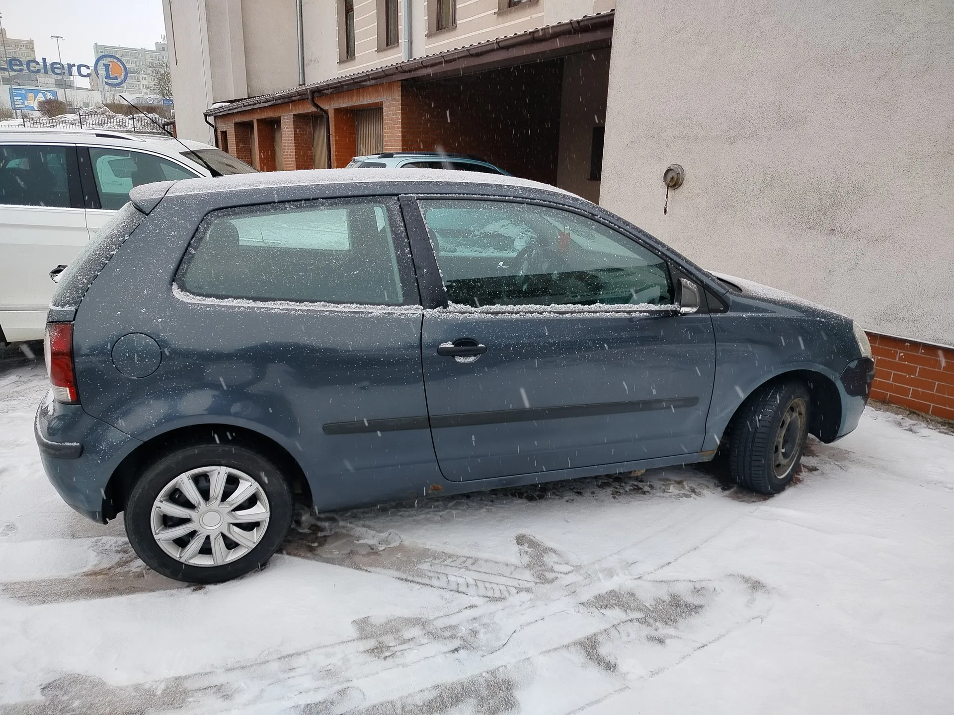 Niebieski samochód osobowy typu hatchback pokryty śniegiem zaparkowany na śnieżnej powierzchni przed budynkiem z ceglaną i beżową elewacją. W tle widoczny biały samochód oraz znak z napisem 'eclerc' i niebieskim logo. Pada śnieg, na ziemi widoczne ślady opon. Budynek posiada kryty wjazd i okna.