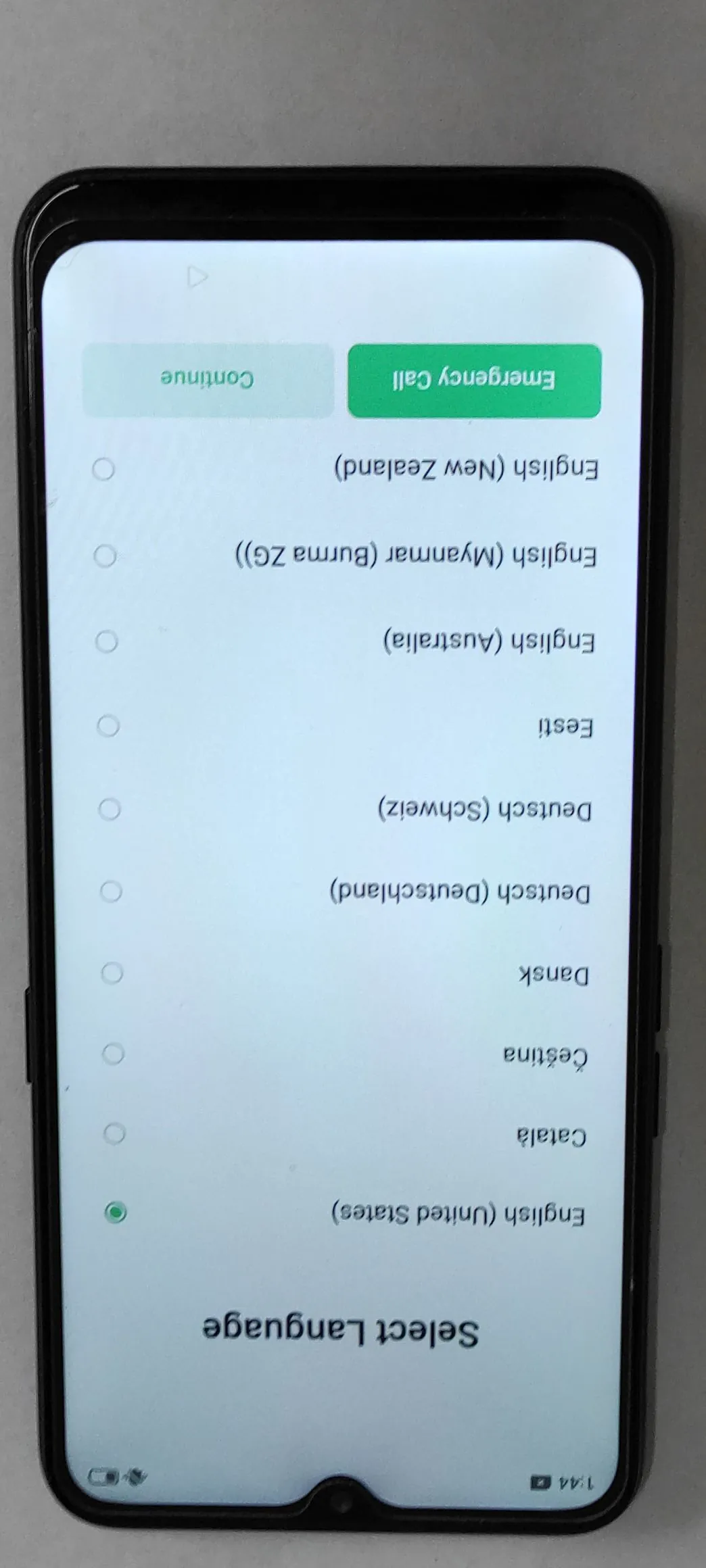 Zdjęcie smartfona wyświetlającego ekran wyboru języka. Na ekranie widnieje napis 'Select Language' oraz lista dostępnych języków, w tym: English (United States), Català, Čeština, Dansk, Deutsch (Deutschland), Deutsch (Schweiz), Eesti, English (Australia), English (Myanmar (Burma ZG)) oraz English (New Zealand). Po prawej stronie znajdują się dwa przyciski: jasnoniebieski 'Continue' oraz zielony 'Emergency Call'. Obraz jest zorientowany poziomo, podczas gdy interfejs telefonu jest pionowy.