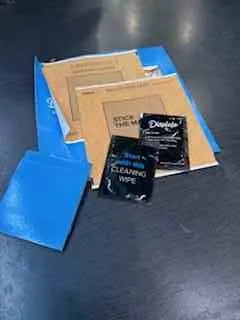 Na ciemnej powierzchni leżą niebieskie i brązowe koperty oraz dwa czarne paczki DHL Express. Na brązowej kopercie widoczny jest napis 'DHL' oraz dodatkowy tekst. Czarne paczki mają wyraźnie widoczne logo 'DHL Express'.