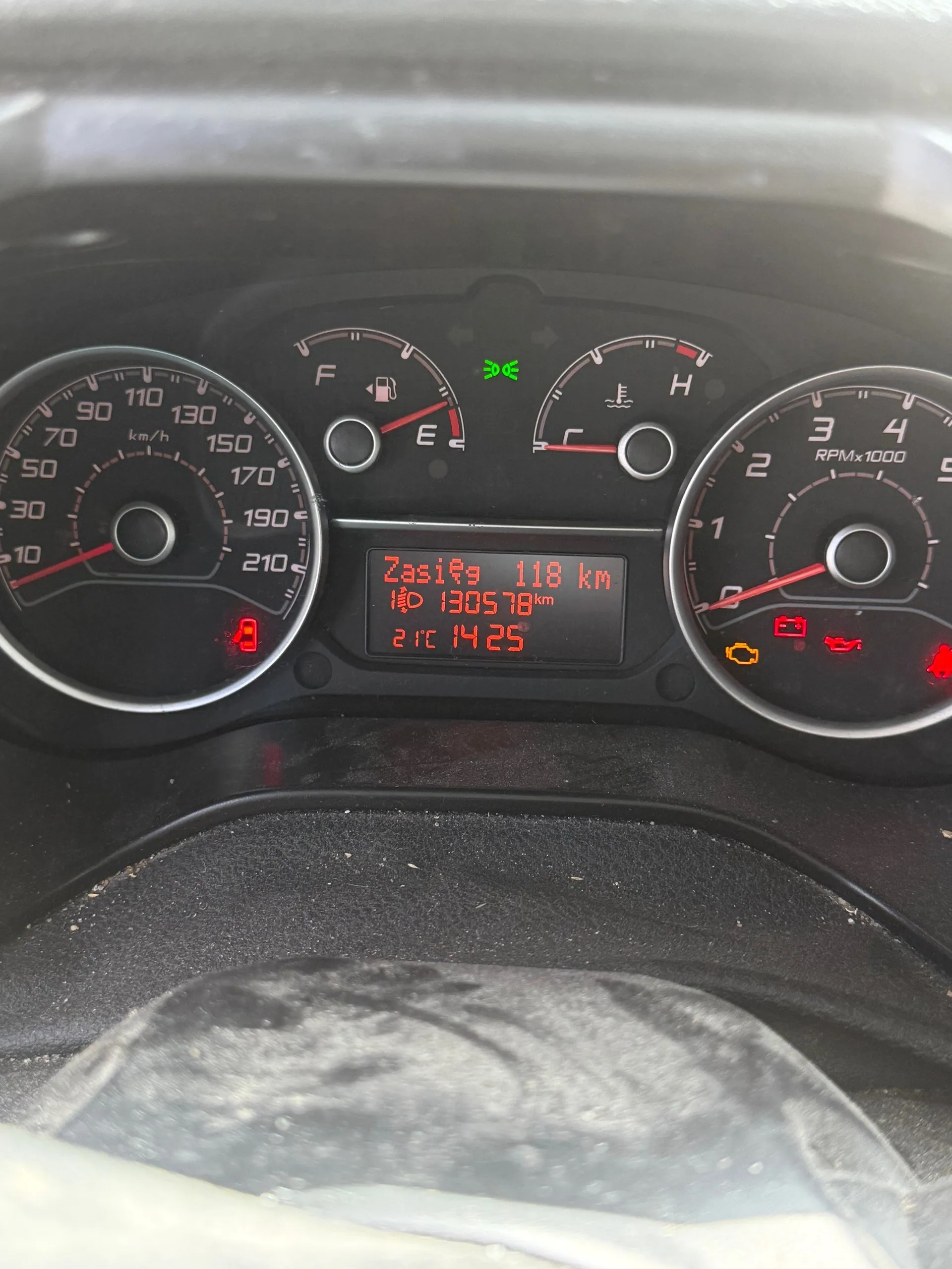 Tablica przyrządów samochodowych z czarnym tłem. Po lewej stronie wskaźnik prędkości do 210 km/h z czerwoną igłą wskazującą około 10 km/h. W centrum wyświetlacz z czerwonym tekstem: "Zasięg 118 km", "130578 km", "21°C 14:25". Nad wyświetlaczem zielony symbol świateł mijania. Po prawej wskaźnik obrotomieru do 5 RPMx1000 z czerwoną igłą wskazującą około 1000 obr./min. Widoczne czerwone lampki ostrzegawcze: akumulator, olej, oraz żółta lampka. Na dole fragment kierownicy z czarną obiciem. Wskaźniki paliwa i temperatury chłodniczy pośrodku, z igłami wskazującymi poziom paliwa blisko E i temperaturę w zakresie normalnym.