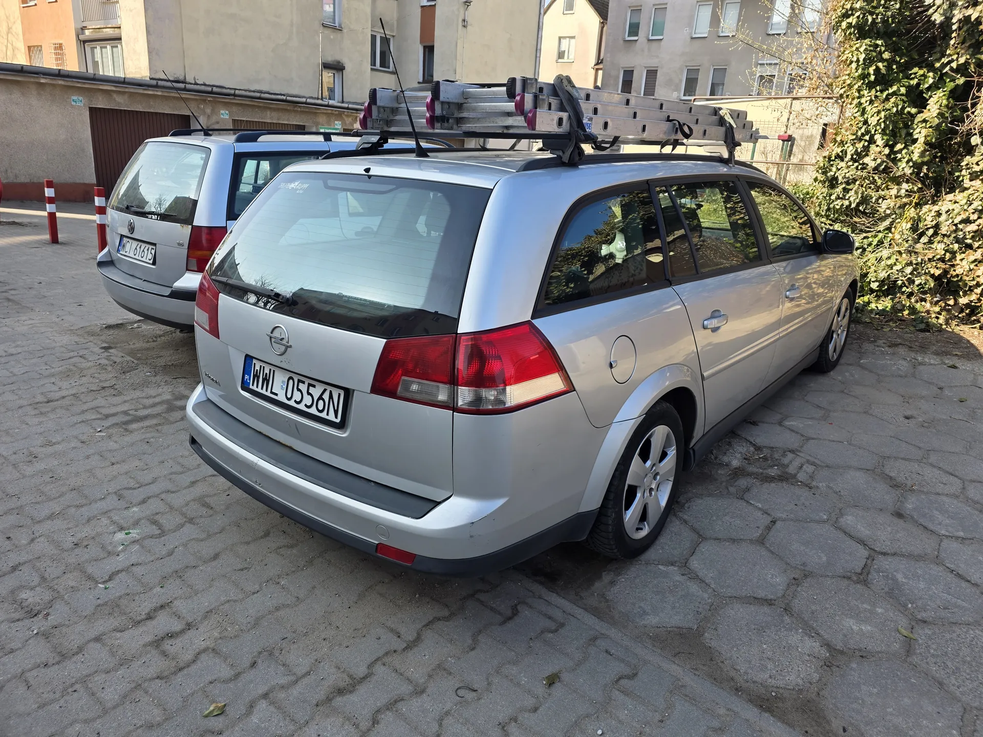 Srebrny samochód typu kombi marki Opel z tablicą rejestracyjną WWL 0556N, na dachu załadowana drabina. W tle drugi srebrny samochód z tablicą WCI 6165, budynki mieszkalne, zielony pnącze oraz czerwono-białe słupki. Samochody zaparkowane na brukowanej powierzchni.