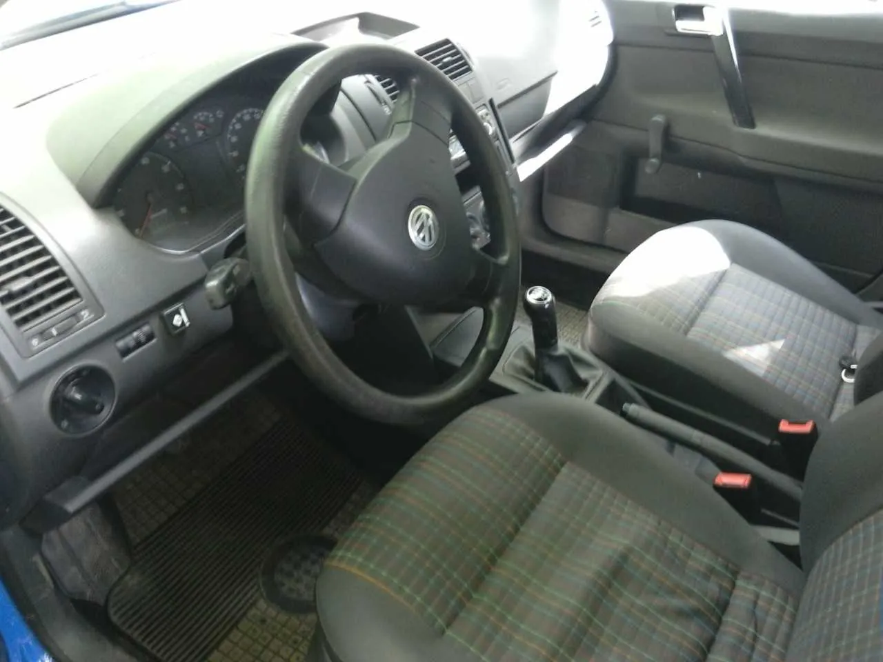 Wnętrze samochodu marki Volkswagen z widocznym kierownicą z logo VW, tablicą rozdzielczą z licznikami, przyciskami klimatyzacji po lewej stronie, dźwignią zmiany biegów oraz siedzeniami w kratę w kolorach szarym, zielonym i brązowym. W tle widoczne są czarne drzwi z uchwytem i oknem.