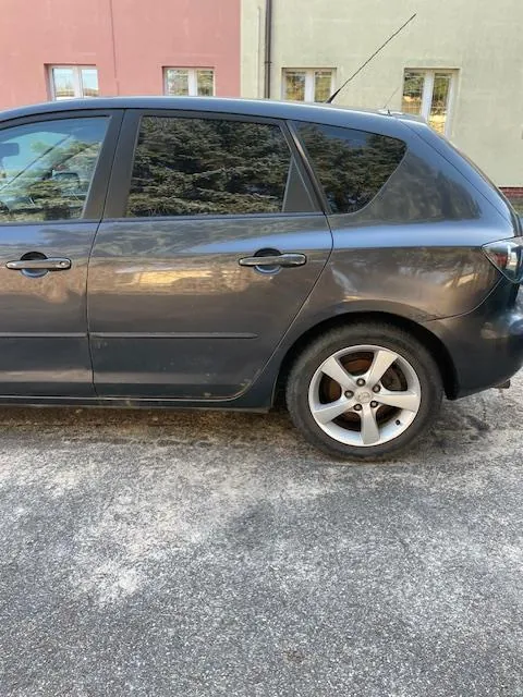 Szary samochód typu hatchback zaparkowany na asfaltowej powierzchni. Widoczna boczna strona pojazdu z czterema drzwiami, srebrnymi felgami i anteną na dachu. W tle budynki: różowy i beżowy z oknami. Asfalt pokryty białymi plamami.