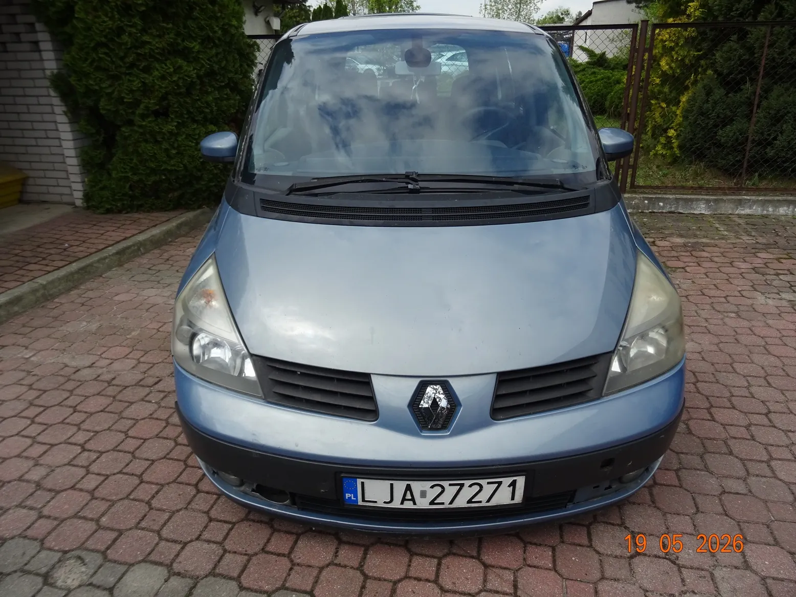 Jasnoniebieski samochód marki Renault zaparkowany przodem do obiektywu na kostce brukowej. Widoczna jest przednia część pojazdu z tablicą rejestracyjną o numerze LJA 27271. W tle znajdują się zielone krzewy i fragment białego ogrodzenia.