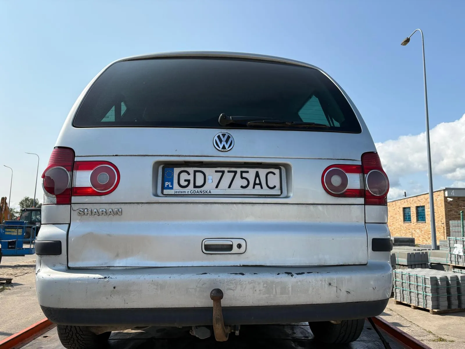 Tył srebrnego samochodu Volkswagen Sharan z tablicą rejestracyjną GD 775AC. Na tablicy widoczne są napisy 'PL' oraz 'jestem z GDANSKA'. Samochód stoi na torach, w tle budynek z cegły, latarnia uliczna, stosy betonowych elementów oraz jasne niebo z kilkoma chmurami. Na bagażniku widoczny jest emblemat VW i napis 'SHARAN'.