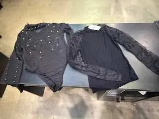 Dwa czarne bluzki z długim rękawem leżące na ciemnym stole. Bluzka po lewej stronie zdobiona białymi kropkami lub naszywkami, po prawej z koronkowym wzorem na rękawach. W tle betonowa podłoga i szafka z szufladami. Widoczna biała etykieta na bluzce po prawej.