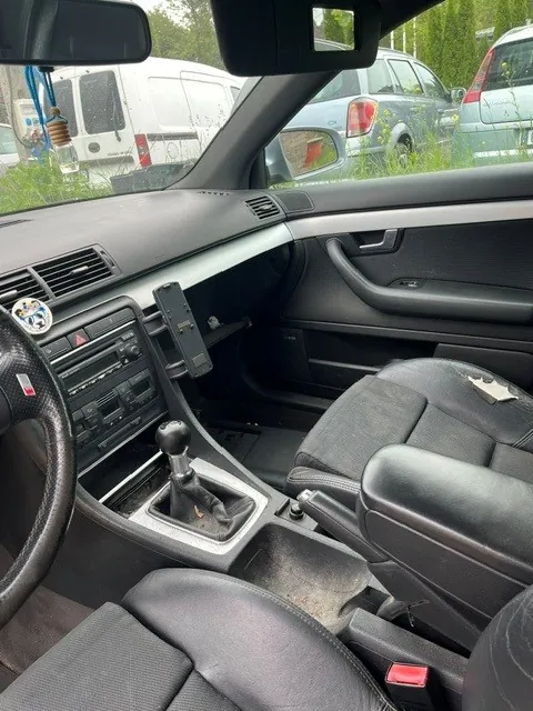 Wnętrze samochodu z perspektywy kierowcy. Widoczna jest czarna skórzana tapicerka foteli, kierownica, konsola środkowa z drążkiem zmiany biegów oraz radio. Do deski rozdzielczej przymocowany jest uchwyt z telefonem komórkowym. Przez przednią szybę i okno pasażera widoczne są inne zaparkowane samochody: biały furgon oraz srebrny samochód osobowy.