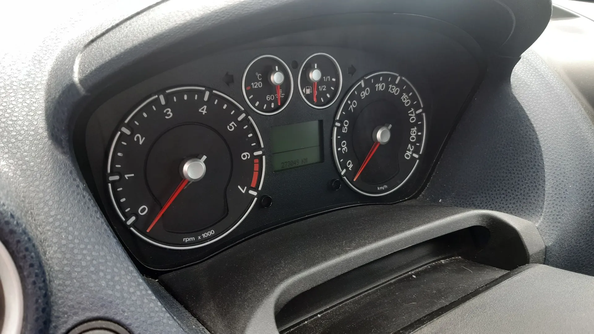 Zbliżenie na deskę rozdzielczą samochodu. Widoczny jest licznik obrotów po lewej stronie z oznaczeniem 'rpm x 1000', prędkościomierz po prawej stronie z oznaczeniem 'km/h', wskaźnik temperatury cieczy chłodzącej oraz wskaźnik poziomu paliwa na górze. W centrum znajduje się mały cyfrowy wyświetlacz z przebiegiem '273049 km'.