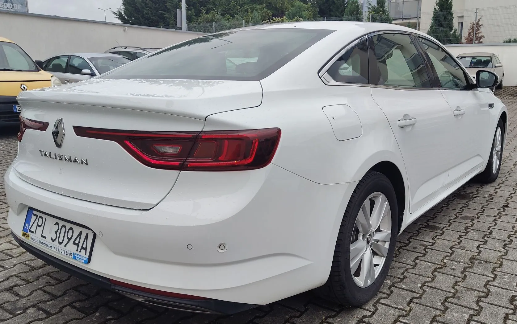 Biały samochód marki Renault Talisman zaparkowany tyłem na kostce brukowej. Na klapie bagażnika widoczne jest logo Renault oraz napis TALISMAN. Samochód posiada polską tablicę rejestracyjną o numerze ZPL 3094A. W tle widoczne są inne zaparkowane samochody oraz budynek.