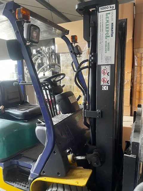 Wózek widłowy o kolorze niebieskim i żółtym z czarnym siedzeniem i kierownicą. Na maszcie widoczne tablice z napisami: '1000 kg', 'KOMATSU', 'LPG', symbolami ostrzegawczymi oraz znakiem CE. W tle pudełka i przemysłowe urządzenia.