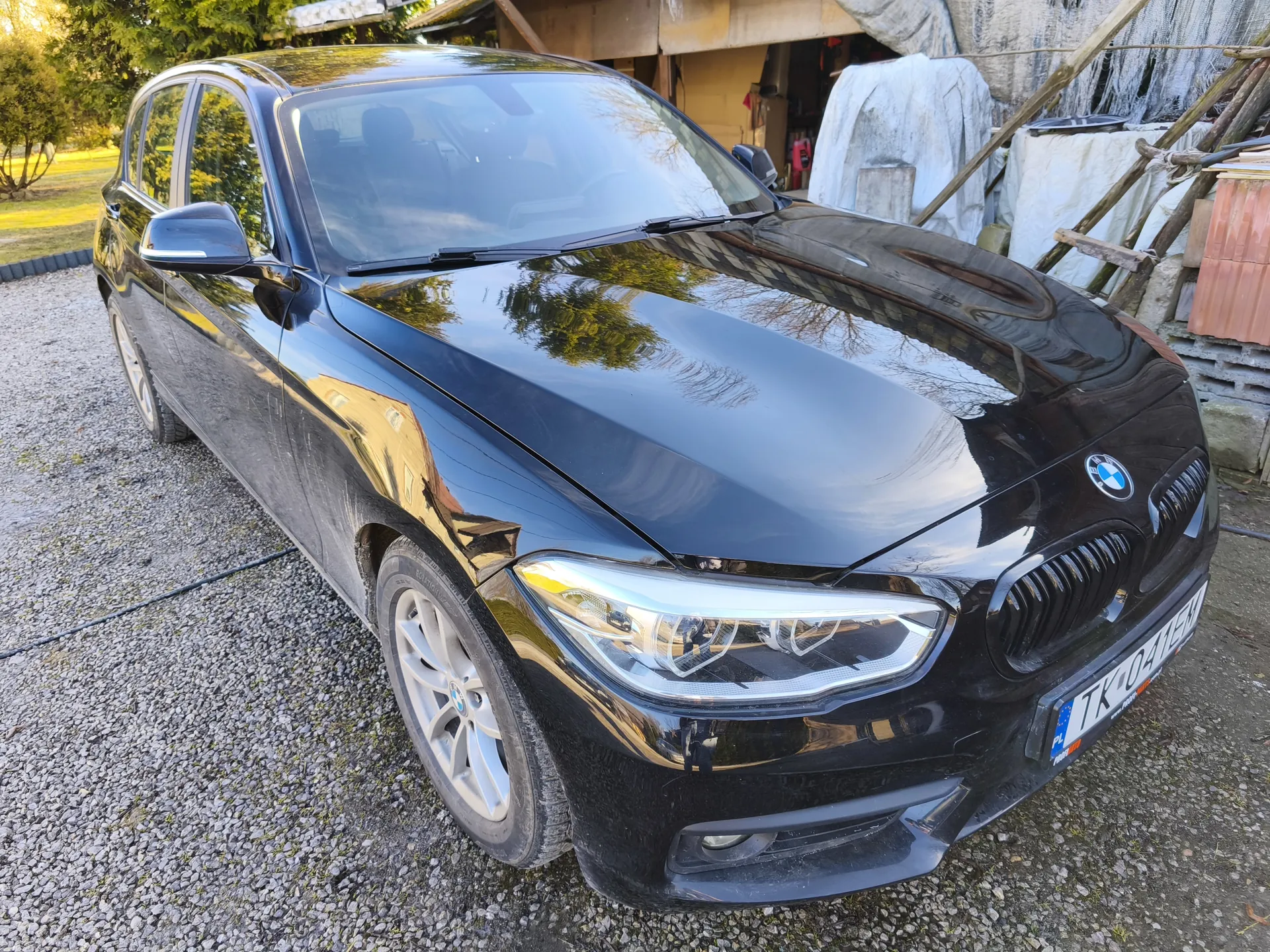 Czarny samochód marki BMW z widocznym logo na masce i tablicą rejestracyjną TK-0415H z polskim flagą. Pojazd stoi na żwirowym podjeździe przed garażem z drewnianym daszkiem. W tle widoczne są zasłonięte przedmioty, drzewa i zielona trawa. Odbicia na błyszczącej karoserii wskazują na słoneczny dzień.