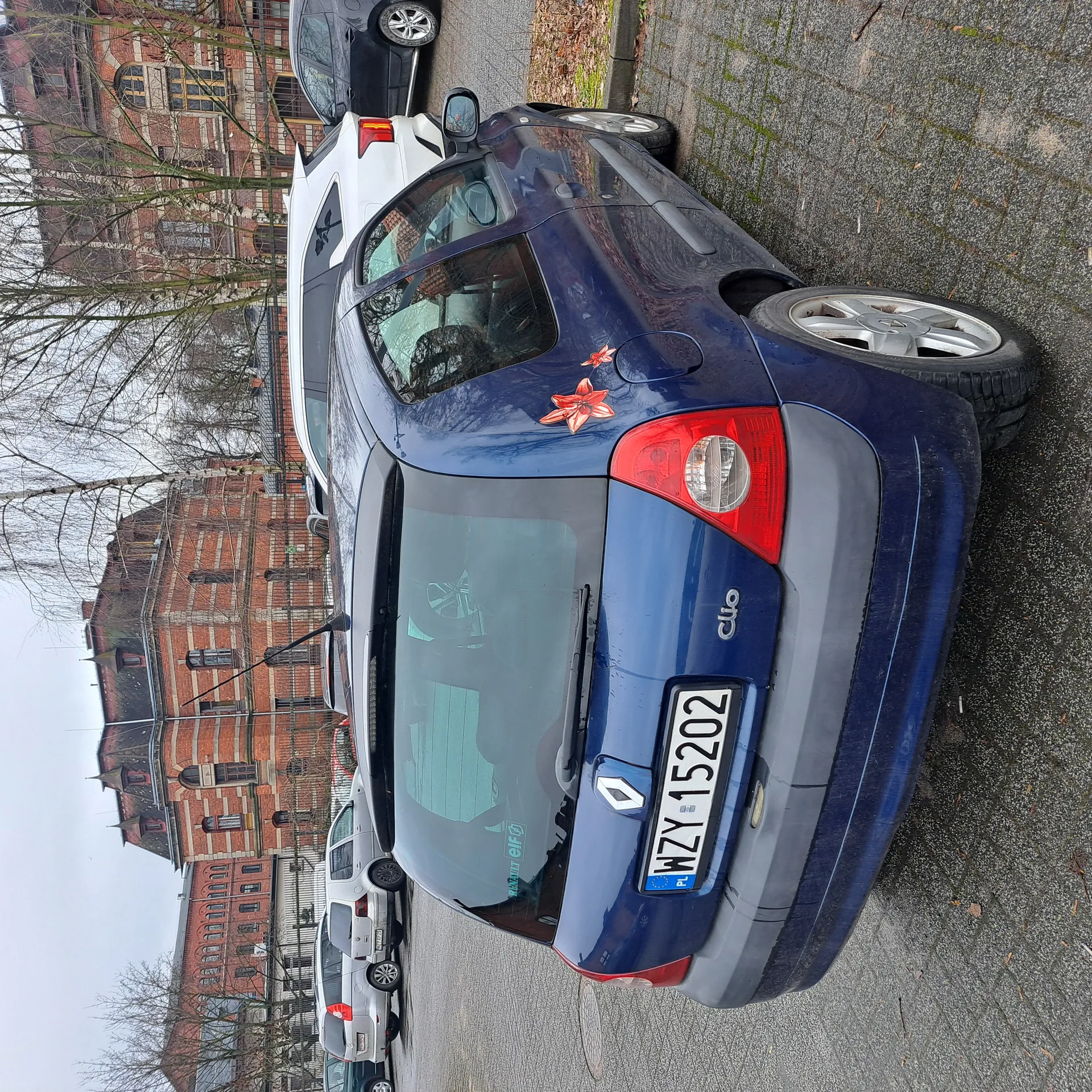 Niebieski samochód Renault Clio zaparkowany na placu parkingowym. Widok od tyłu z tablicą rejestracyjną WZY 15202 (PL). Na tylnej szybie naklejka z napisem 'elf' oraz czerwone kwiatowe naklejki na boku. W tle inne zaparkowane pojazdy, ceglane budynki i drzewa bez liści. Niebo zachmurzone.