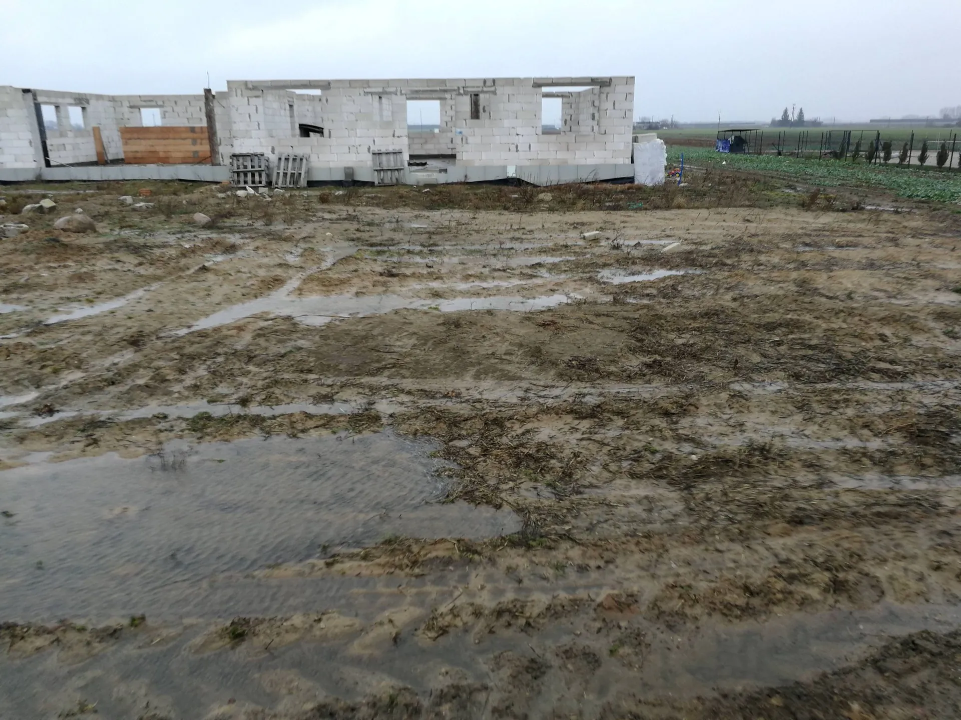 Zdjęcie przedstawia plac budowy w pochmurny dzień. W tle widoczne są nieukończone ściany budynku wzniesionego z białych bloczków betonowych z otworami na okna i drzwi. Przed budynkiem znajduje się rozległy, błotnisty teren z licznymi koleinami i kałużami wody. Po prawej stronie w oddali widać ogrodzenie oraz fragmenty zieleni.