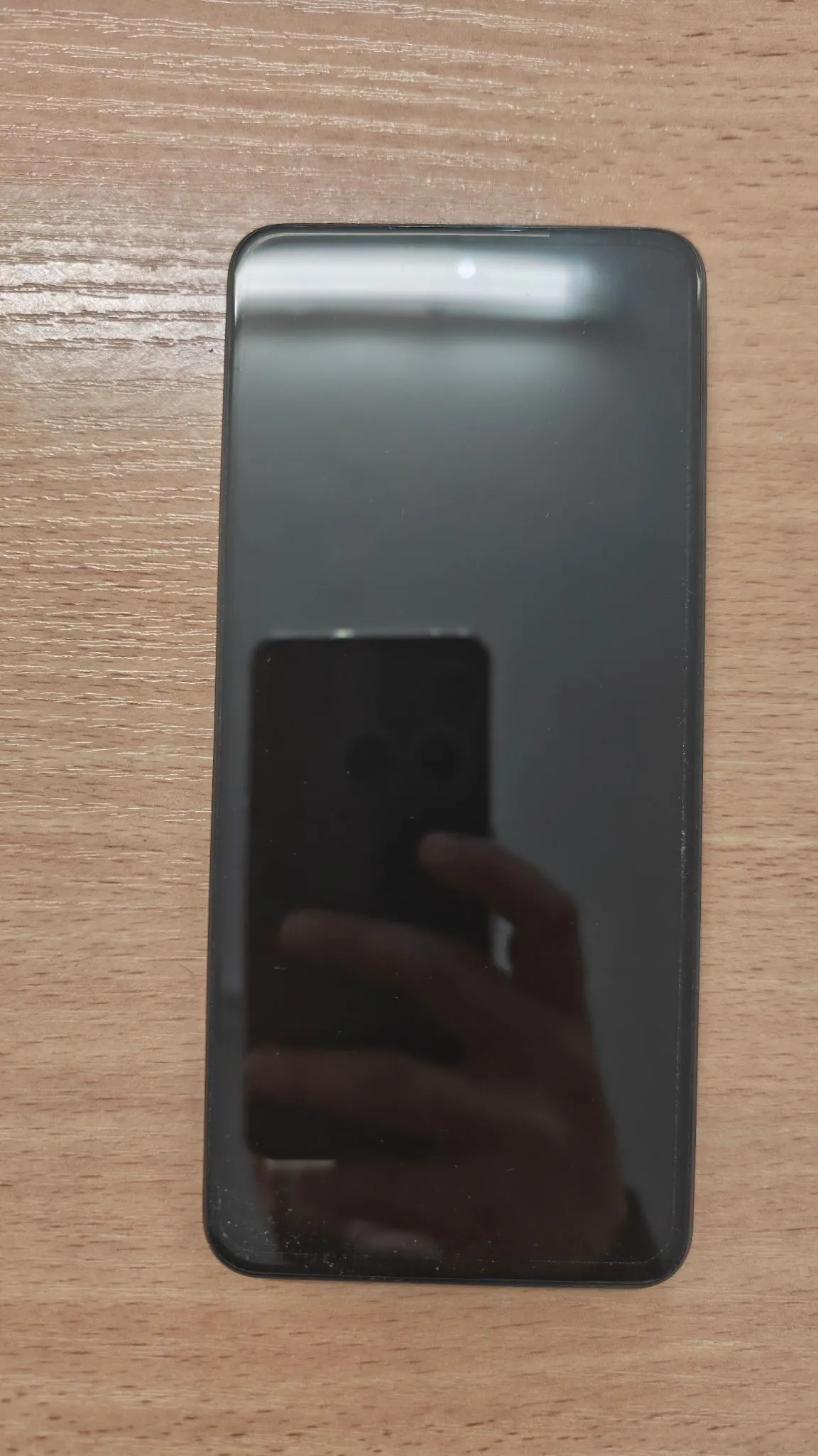 Czarny smartfon z wyłączonym ekranem leżący pionowo na drewnianym blacie. W ciemnej tafli ekranu widoczne jest odbicie osoby trzymającej telefon oraz drugiego urządzenia.
