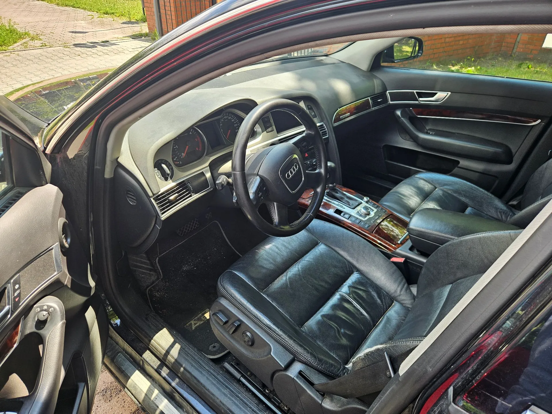 Wnętrze samochodu marki Audi widziane z otwartych drzwi kierowcy. Widoczna jest czarna skórzana tapicerka foteli, czarna kierownica z logo Audi, deska rozdzielcza z analogowymi zegarami oraz konsola środkowa z wykończeniem w kolorze ciemnego drewna. Samochód ma lewostronną kierownicę.