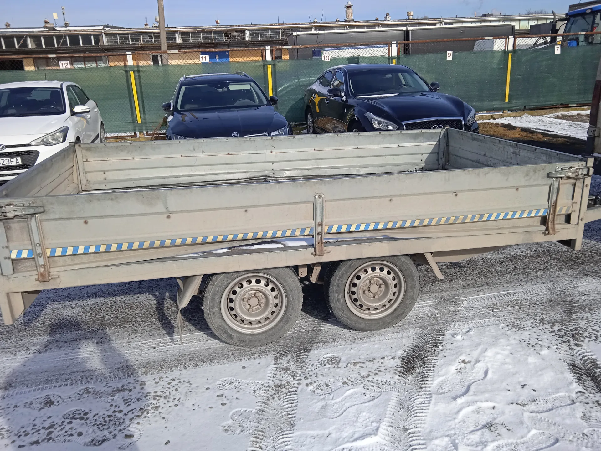 Na pierwszym planie metalowy przyczepa z niebiesko-żółtymi paskami, z dwoma kołami i śladami opon na śnieżnej nawierzchni. W tle trzy samochody: biały po lewej stronie z tablicą rejestracyjną 23FA, ciemnoniebieski w środku i czarny Infiniti po prawej. Za nimi zielone ogrodzenie z żółtymi słupkami i tabliczkami z numerami 12, 11, 10, 9, 7. W oddali budynki przemysłowe i jasne niebo. Śnieg pokrywa część terenu, widoczne ślady opon.