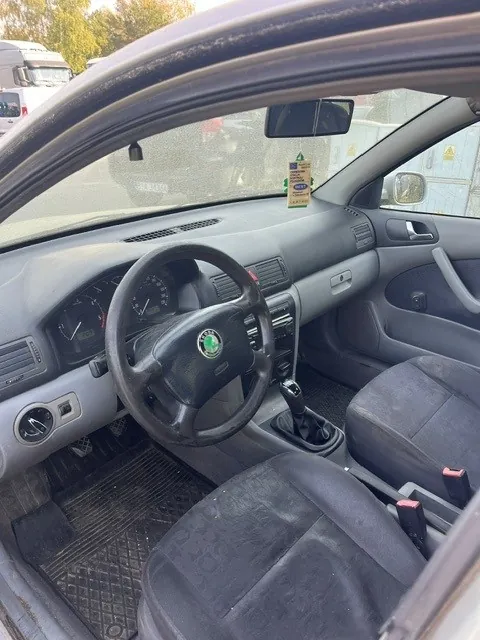 Wnętrze samochodu marki Skoda widziane z perspektywy pasażera. Widoczna jest szara deska rozdzielcza, czarna kierownica z logo marki, fotele z ciemnej tkaniny oraz lewarek zmiany biegów. Na lusterku wstecznym wisi żółta zawieszka. Przez przednią szybę widoczne są inne zaparkowane pojazdy.