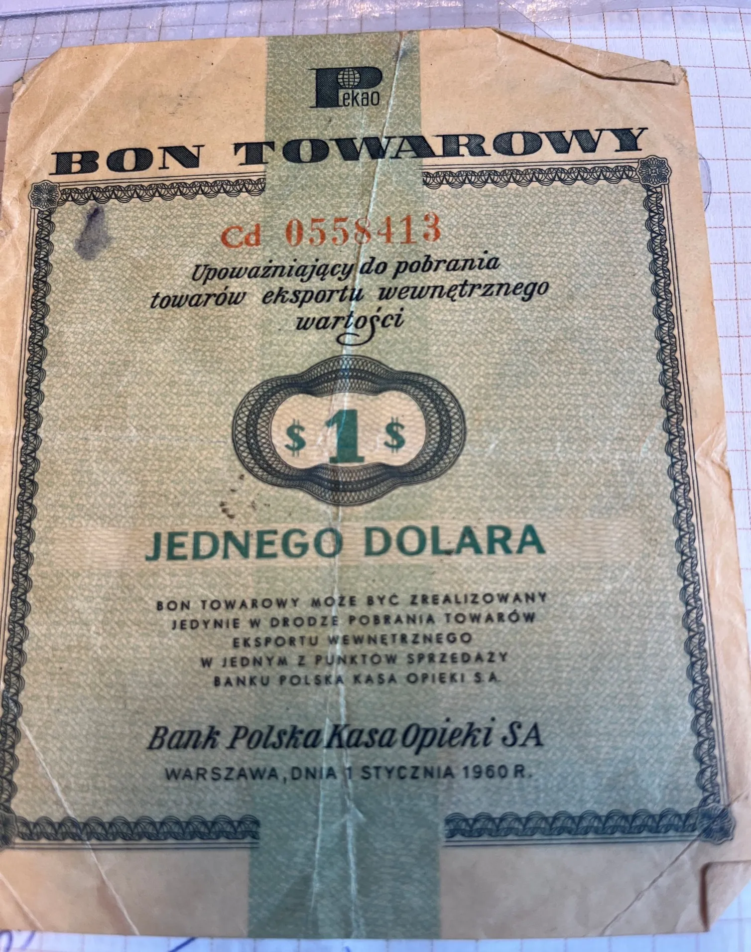 Stary, pożółkły bon towarowy z logo PKO (Bank Polska Kasa Opieki). Na górze widnieje napis 'BON TOWAROWY'. Poniżej znajduje się czerwony numer seryjny 'cd 0558413' oraz tekst: 'Upoważniający do pobrania towarów eksportu wewnętrznego wartości'. W centralnej części znajduje się ozdobny owal z napisem '$ 1 $', a pod nim duży napis 'JEDNEGO DOLARA'. W dolnej części znajduje się mniejszy tekst informujący, że bon może być zrealizowany w punktach sprzedaży Banku Polska Kasa Opieki S.A., oraz podpis 'Bank Polska Kasa Opieki SA' i data 'WARSZAWA, DNIA 1 STYCZNIA 1960 R.'. Całość otoczona jest ozdobną ramką.