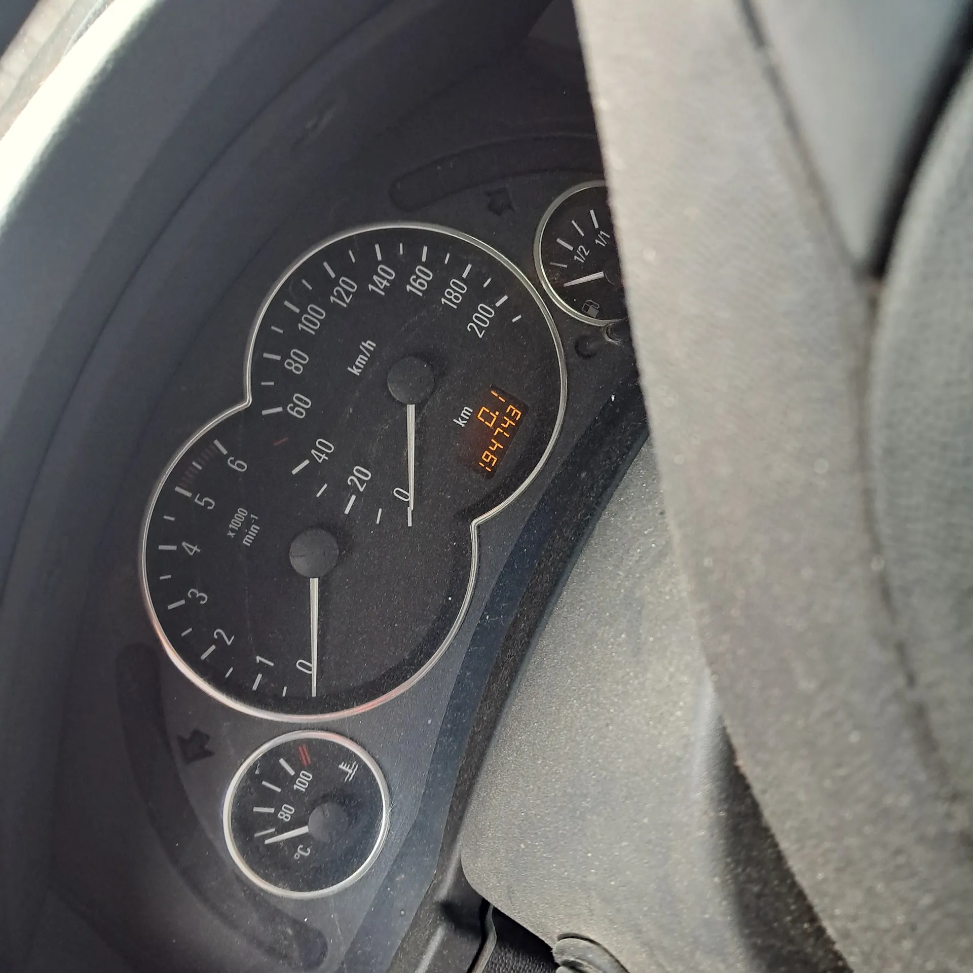 Zbliżenie na deski rozdzielcze samochodu z licznikiem prędkości (0-200 km/h), tachometrem (0-6 x1000 obr./min), wskaźnikiem temperatury chłodniczki (0-120°C), wskaźnikiem paliwa oraz cyfrowym wyświetlaczem pokazującym przebieg 19473 km. Widoczny jest symbol ostrzegawczy przedstawiający samochód z wykrzyknikiem. Tło stanowi czarna deska rozdzielcza z białymi oznaczeniami i częściowo widoczna kierownica.