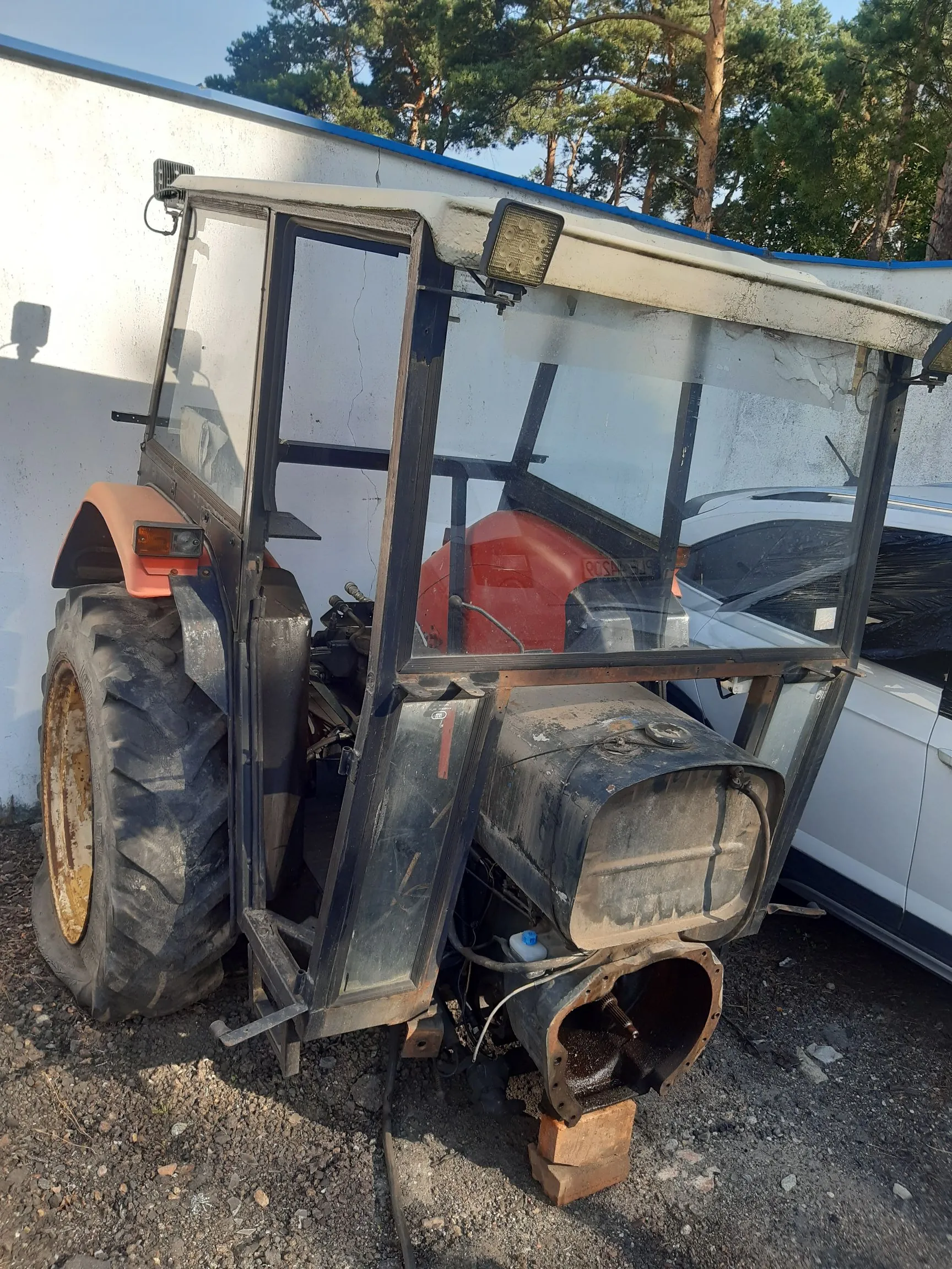 Zdjęcie przedstawiające fragment czerwonego ciągnika rolniczego z zamontowaną szklaną kabiną. Maszyna stoi na piaszczystym podłożu obok białego budynku i zaparkowanego białego samochodu. Widoczne jest duże tylne koło z wyraźnym bieżnikiem oraz konstrukcja kabiny z ramami i szybami.