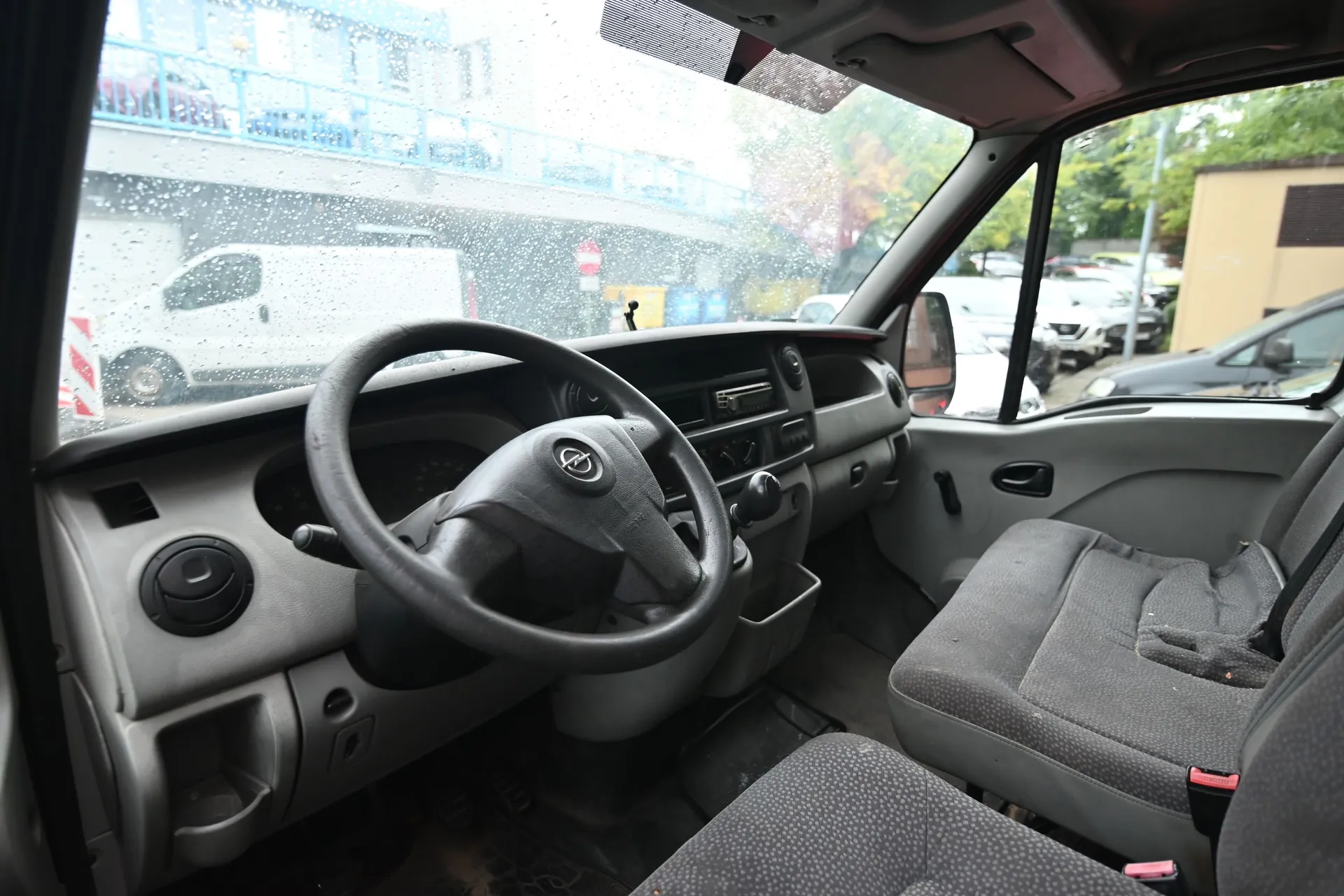Wnętrze kabiny samochodu dostawczego marki Opel, widok z perspektywy pasażera. Widoczna jest szara deska rozdzielcza, kierownica z logo producenta, dwa szare fotele z materiału oraz przednia szyba pokryta kroplami deszczu. Przez szybę widać biały samochód dostawczy zaparkowany na zewnątrz oraz fragmenty budynków.