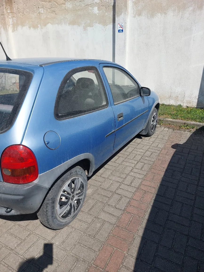 Niebieski samochód osobowy typu hatchback zaparkowany na brukowanej nawierzchni. Samochód jest lekko zabrudzony. W tle biała, częściowo obłupana ściana z niebieskim znakiem drogowym. Na nawierzchni widoczny cień osoby fotografującej oraz pas trawy przy ścianie.