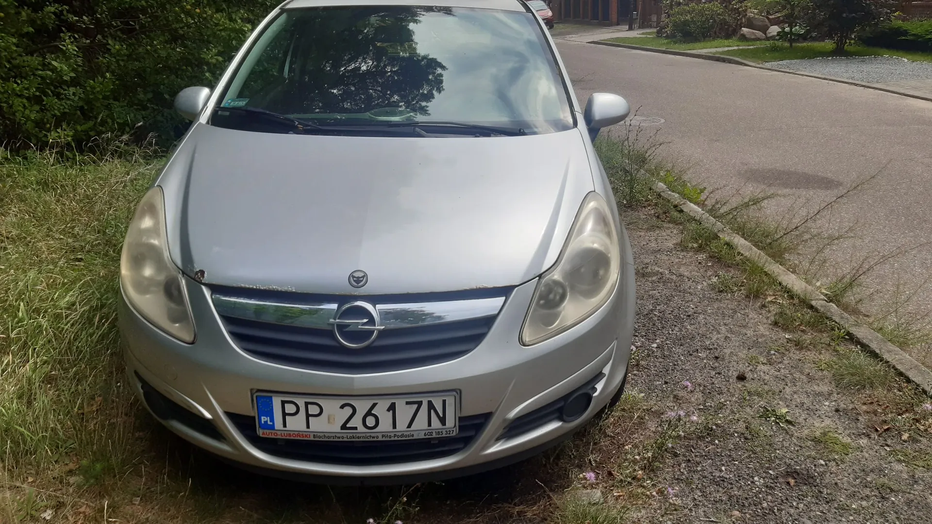 Srebrny samochód marki Opel zaparkowany na poboczu drogi, obok trawy i krawężnika. Widoczny jest przód pojazdu z tablicą rejestracyjną o numerze PP 2617N. Poniżej numeru rejestracyjnego znajduje się napis: AUTO-LUBORSKI Błocharstwo-Lakiernictwo Piła-Podlasie 600 185 327.