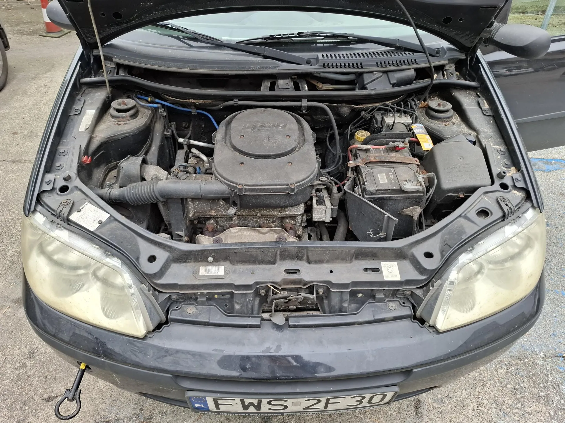 Czarny samochód z otwartą maską, ukazujący komorę silnika. Widoczny silnik z napisem "FIAT" na pokrywie, akumulator z czerwonymi i czarnymi przewodami, różne przewody i węże. Przednia część pojazdu z żółknącymi reflektorami i tablicą rejestracyjną PL FWS 2F30. Tło: betonowa powierzchnia, fragment pomarańczowo-białego słupa ostrzegawczego po lewej stronie.