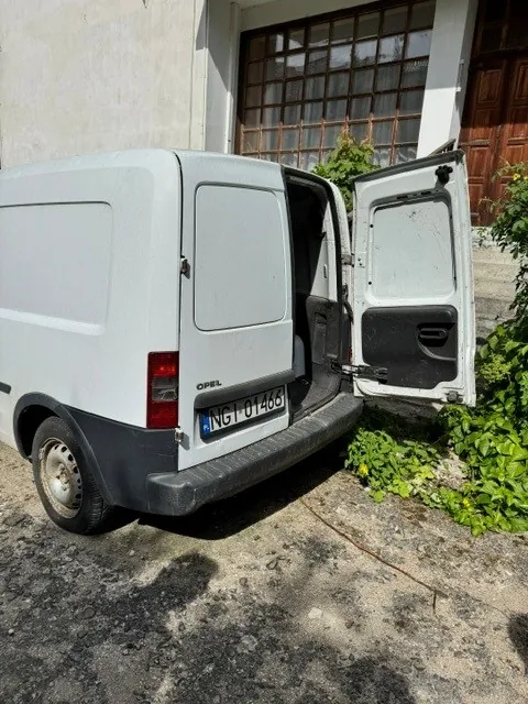 Biały samochód dostawczy marki Opel z otwartymi tylnymi drzwiami, zaparkowany na betonowym podłożu obok budynku. Pojazd posiada tablicę rejestracyjną o numerze NGI 01466. W tle widoczne jest okno z kratami oraz zielone rośliny.